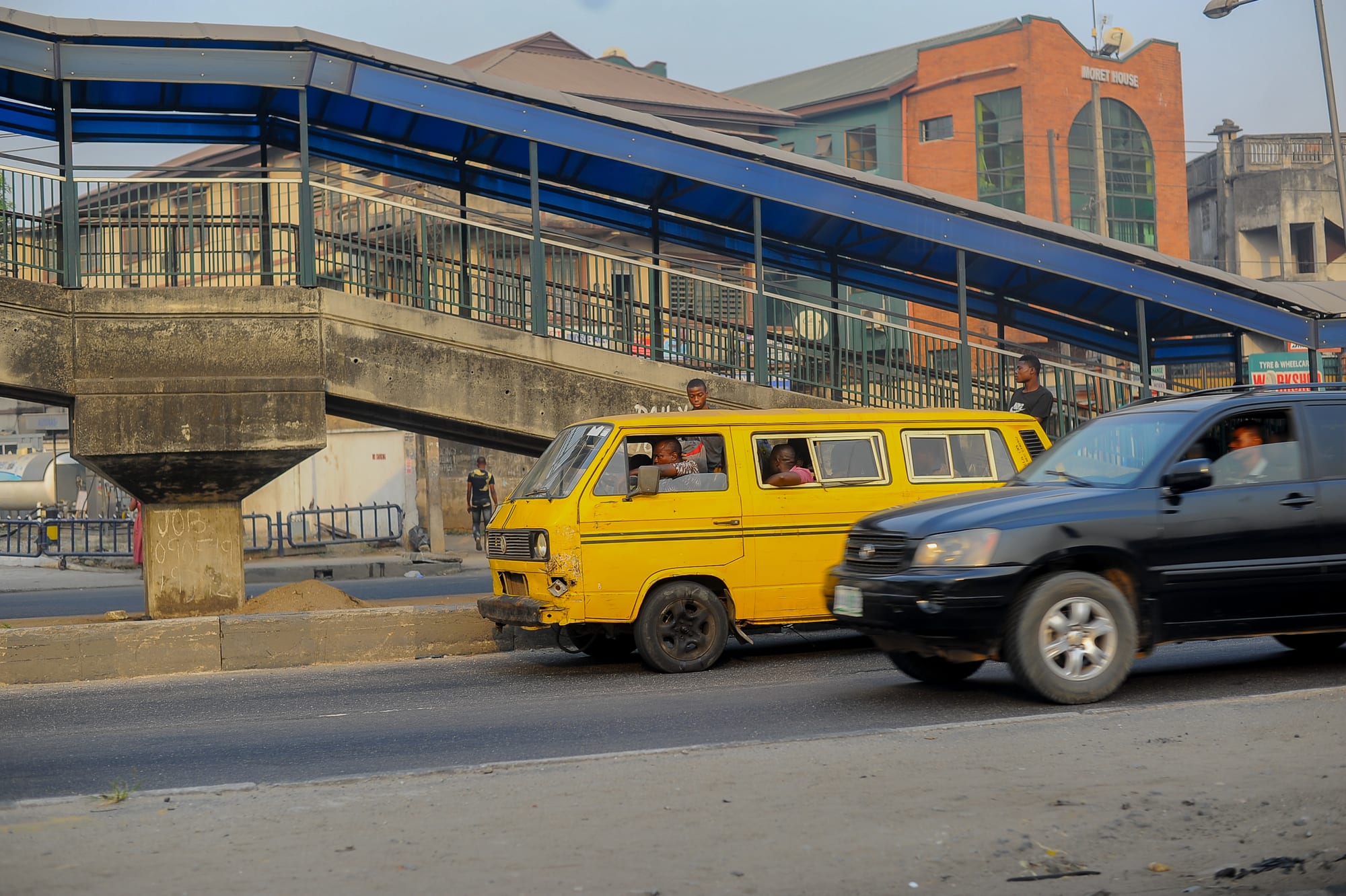 A yellow danfo minibus and a black car passing under a pedestrian footbridge on a busy Lagos street