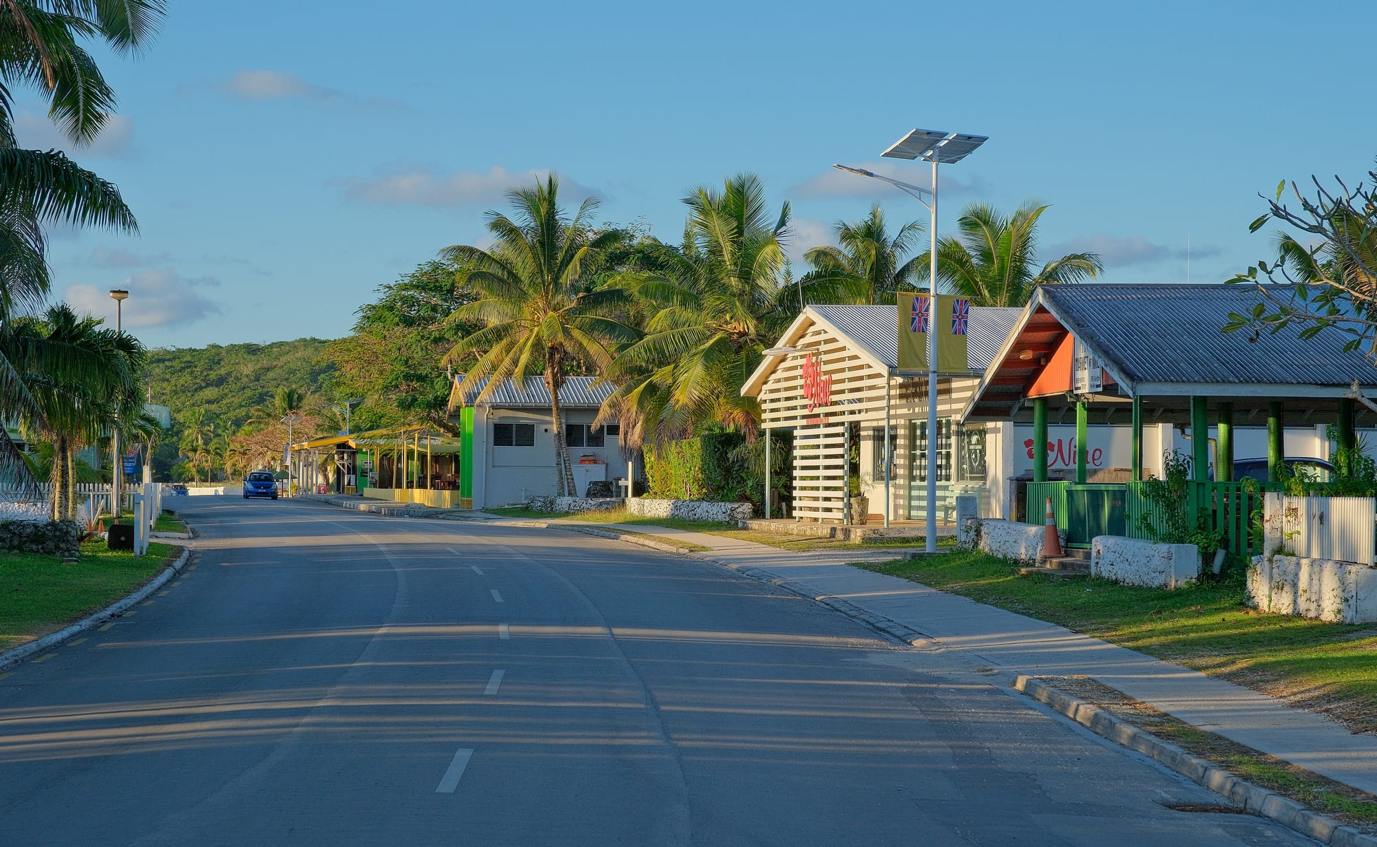 A quiet paved road through Alofi lined with low shops and palm trees under a clear evening sky