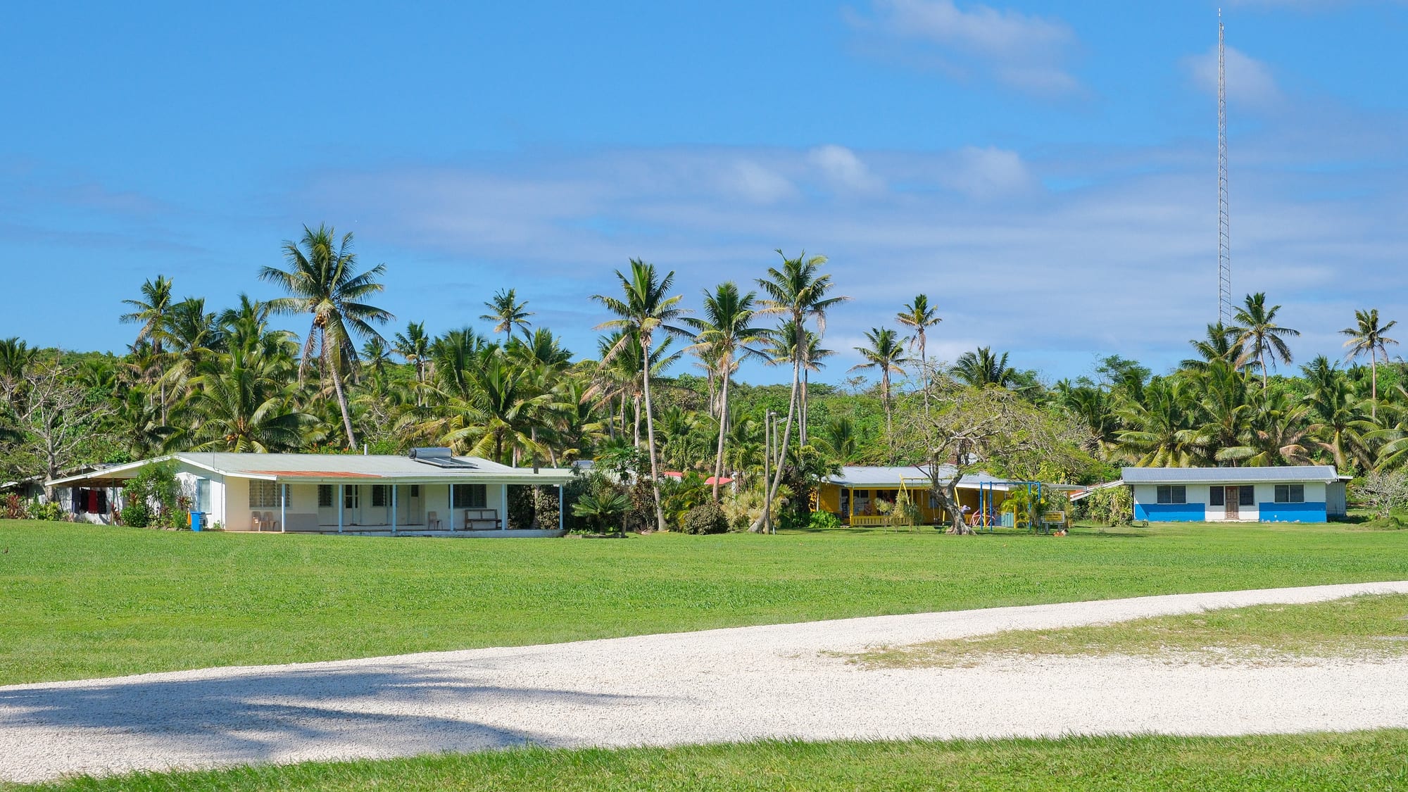 Low single-storey houses and a pre-school spaced across an open grassy area with palm trees behind, in Lakepa village