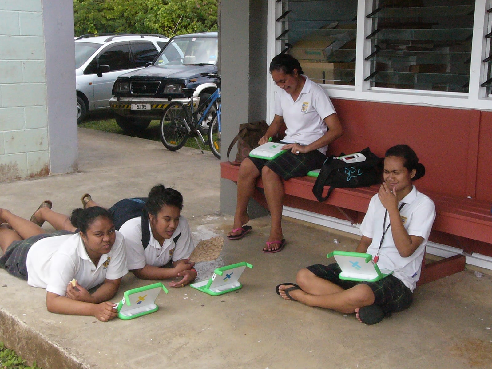 Four teenage girls in school uniform sitting and lying on a veranda floor, each with a small green laptop