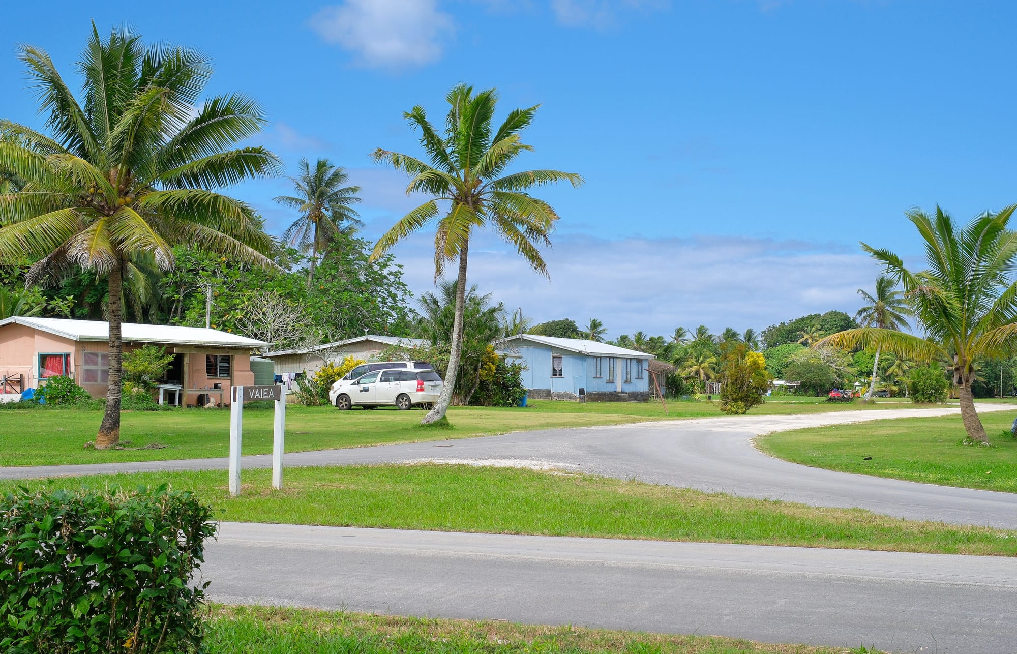 Single-storey houses painted pink and pale blue sit among palm trees and open lawns beside a quiet curving road, with a white sign reading Vaiea