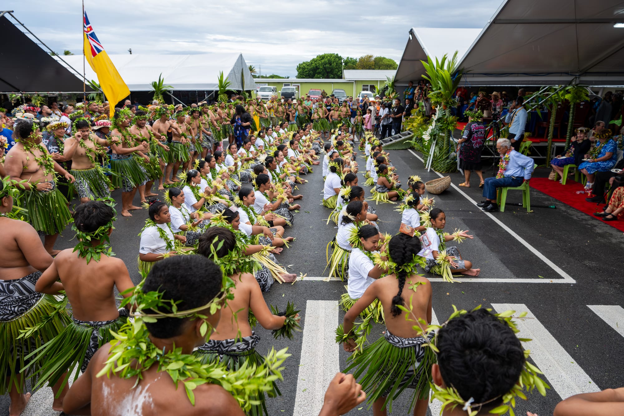 Rows of dancers in leaf skirts and green leaf garlands sit on a paved square performing before guests seated under awnings, with Niue's yellow flag flying on a pole