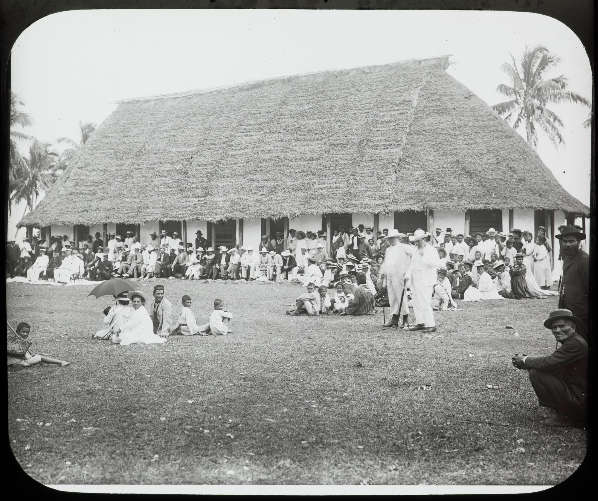 A black and white photograph of a large crowd, many in white clothes and hats, gathered on the grass outside a long church building with a steep thatched roof