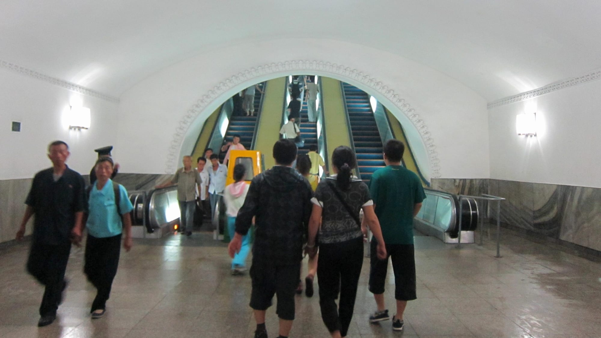 Commuters walking through a station of the Pyongyang Metro, with others riding escalators in the background