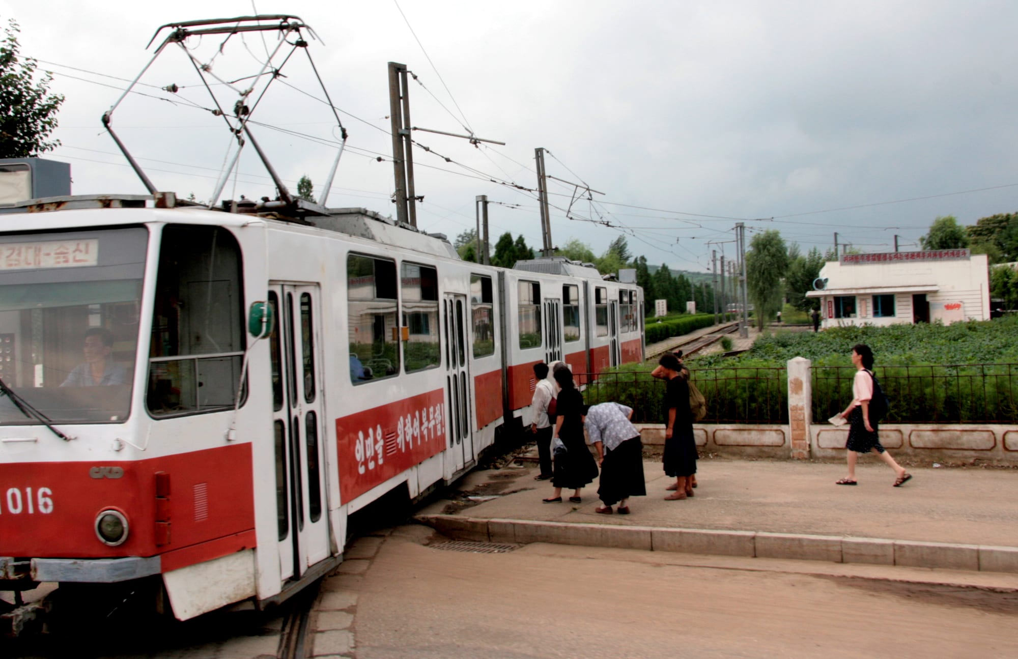 A red-and-white tram stopped at a platform while several people board and a woman walks past on the pavement
