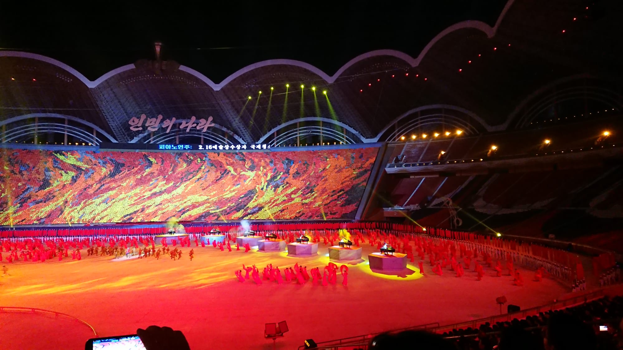 A floodlit stadium at night, with a vast card-section backdrop in red, orange and yellow across the far stands and performers in red spread across the floor below