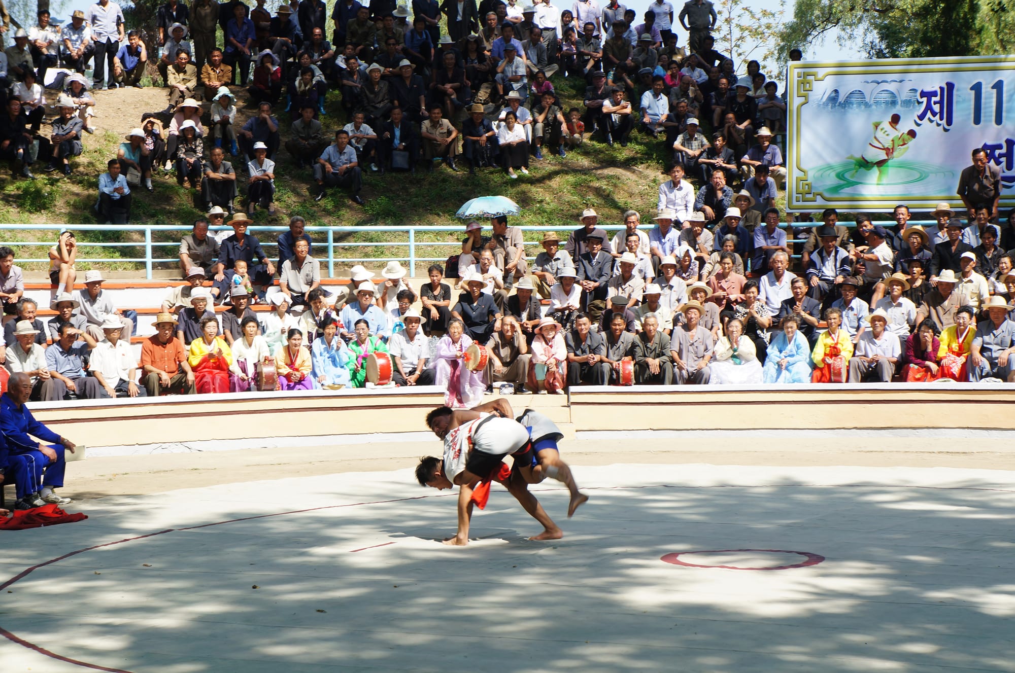 Two wrestlers grapple on a sandy ring, one lifting the other, while a large crowd watches from tiered seating and a grassy bank behind