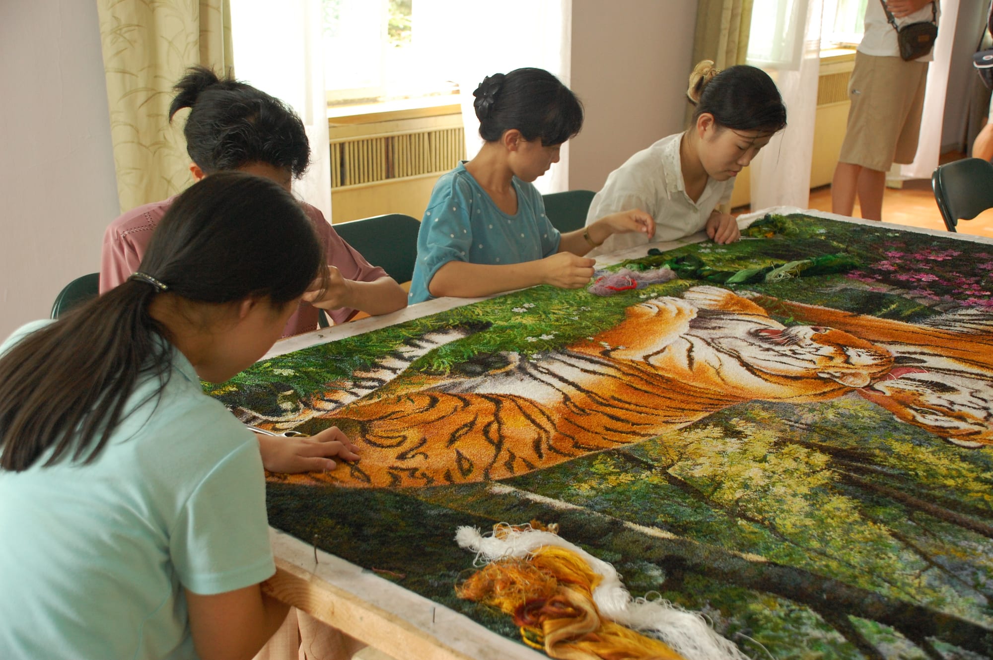 Four women seated around a large frame work with coloured threads on an embroidered picture of a tiger lying in green grass