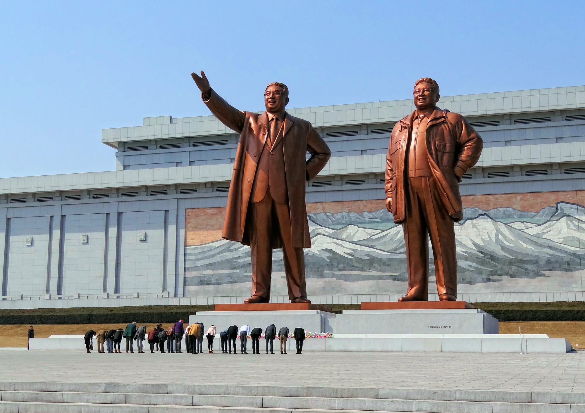A line of visitors bowing in front of two giant bronze statues of North Korea's first two leaders, with a large mosaic of snow-capped mountains on the building behind