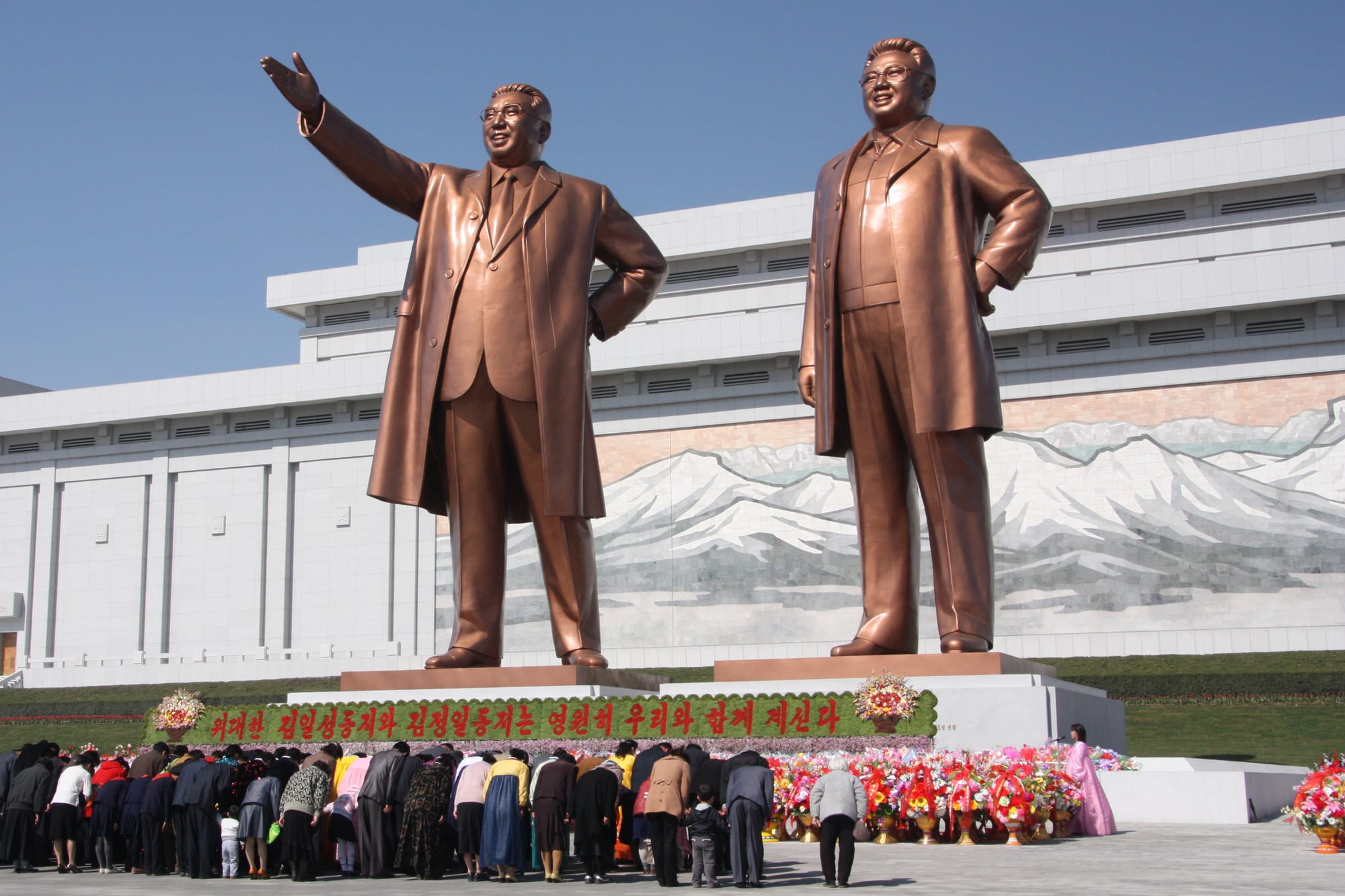 Two large bronze statues of Kim Il Sung and Kim Jong Il on a hillside in Pyongyang, with visitors bowing