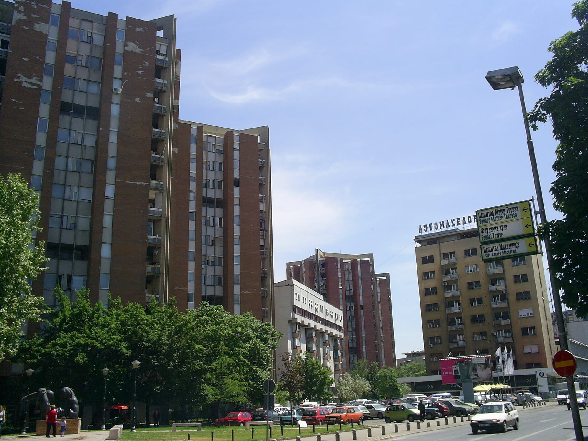 Tall brown and beige apartment blocks along a Skopje street, with parked cars and trees in front