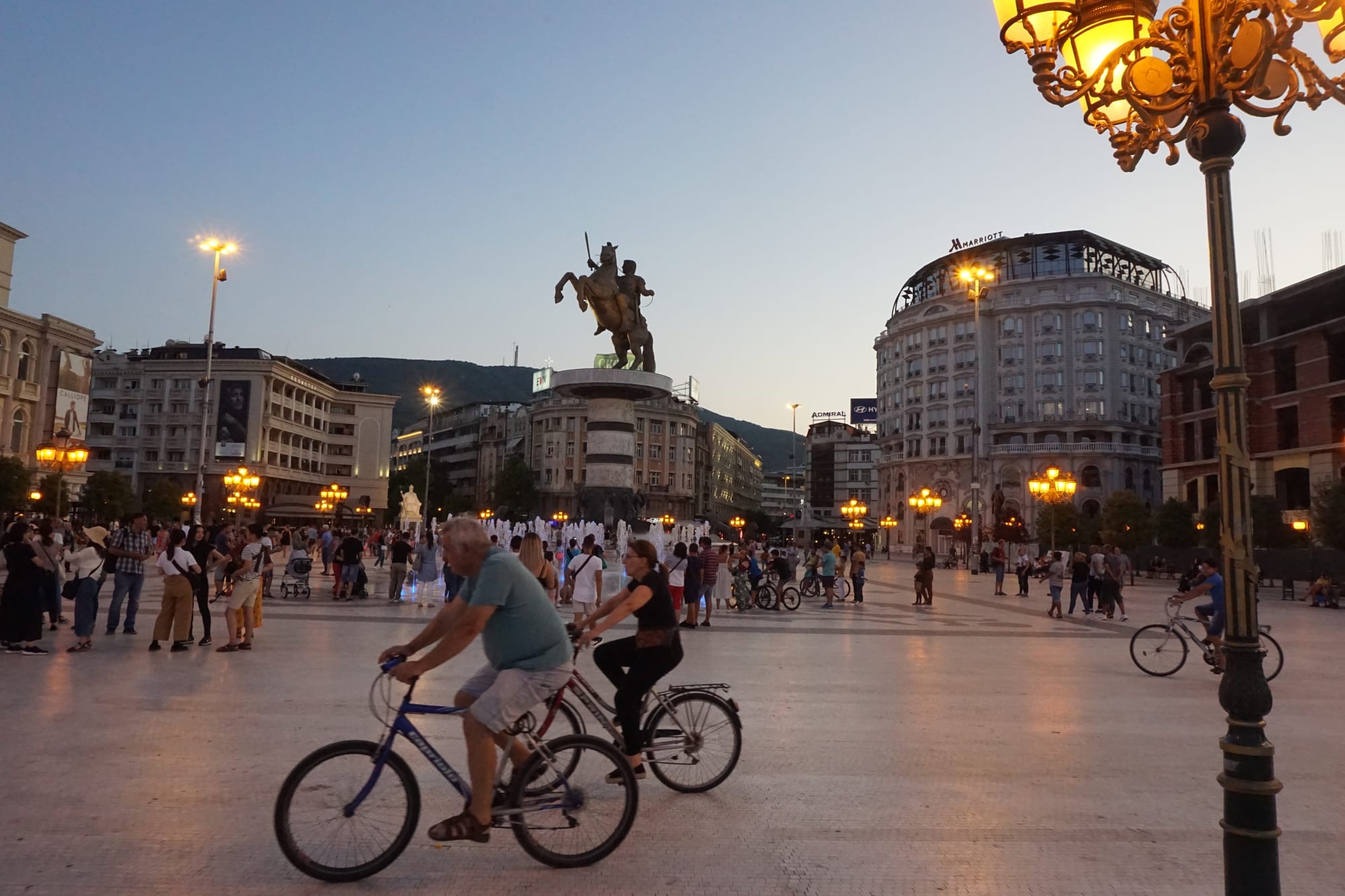 A busy city square at dusk with people cycling, walking and gathered near a lit fountain and an equestrian statue