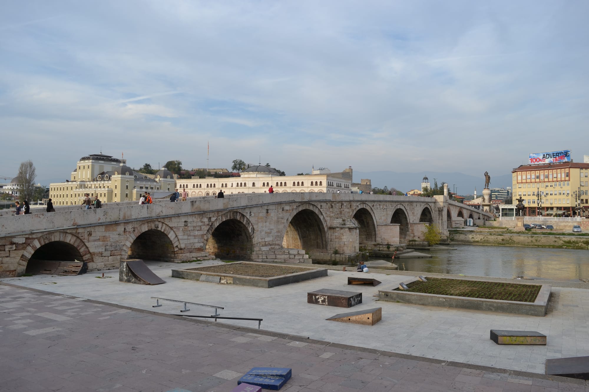 A long stone arched bridge crossing a river, with a mosque and fortress visible behind