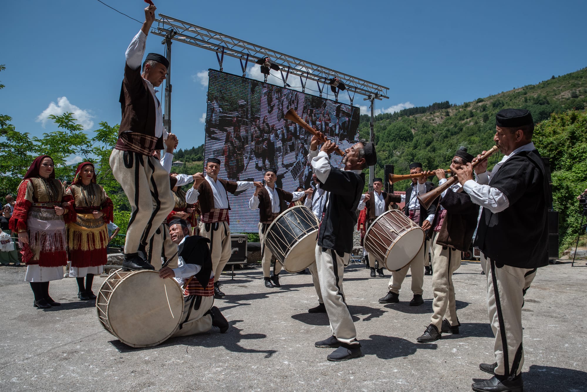 Men in white-and-black folk costume play long wooden shawms and large double-headed drums outdoors, one dancer standing on top of a drum while women in embroidered red dresses watch