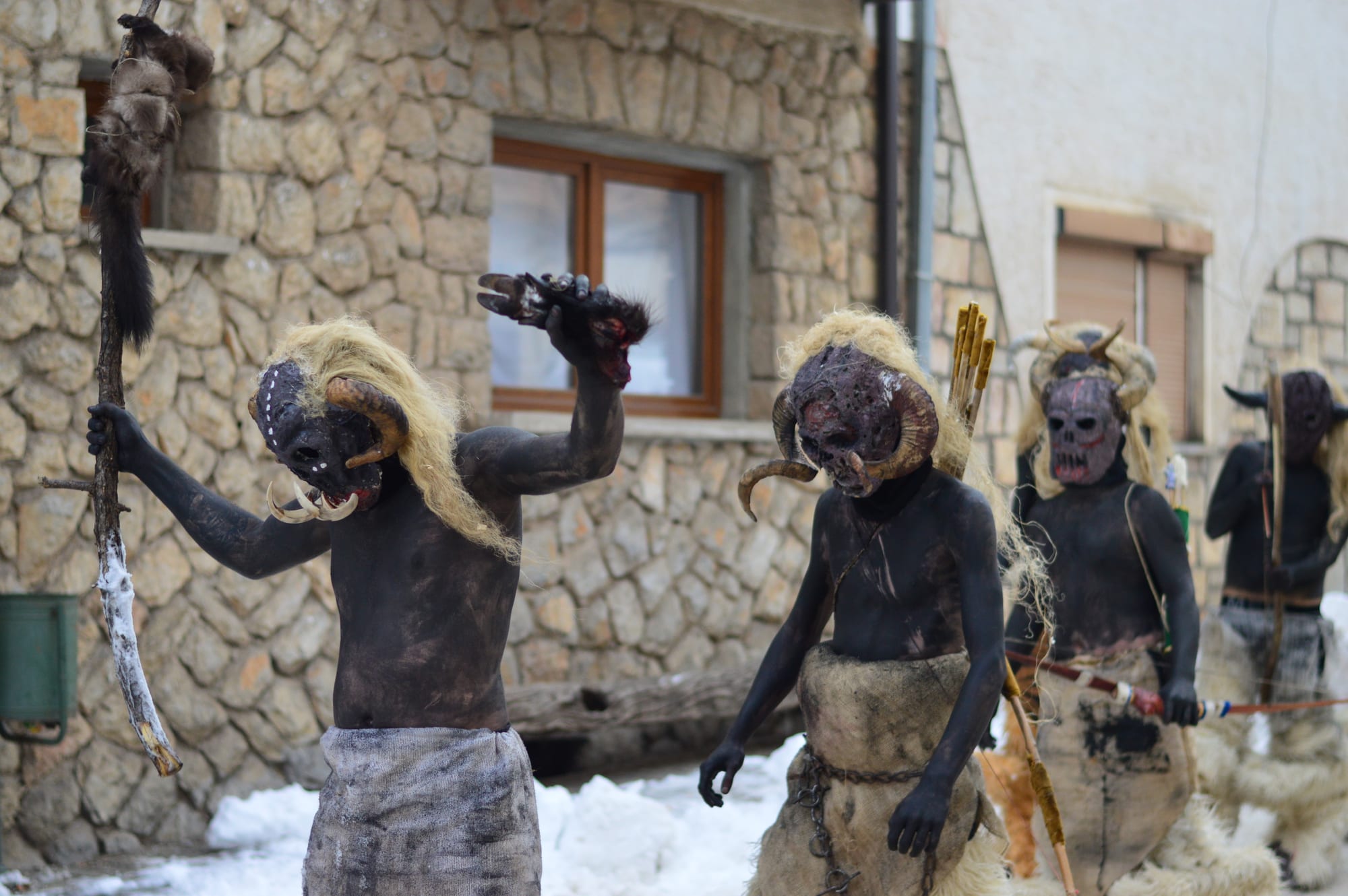 Carnival participants with bodies painted black, wearing horned devil masks, fur and blond wigs and carrying staffs, walk past stone houses in the snow