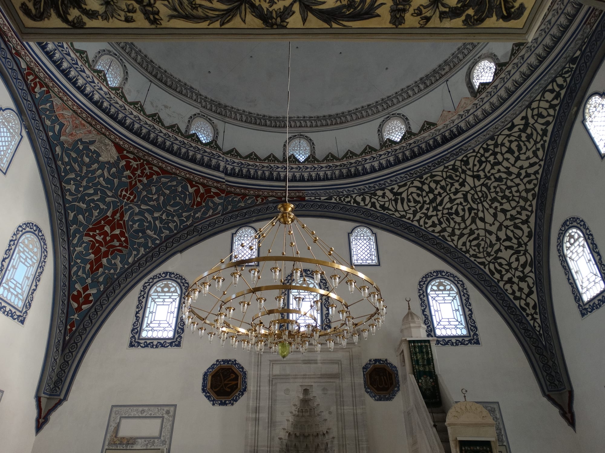 The interior of a mosque with a painted dome, walls decorated with blue and red floral patterns, arched windows, a large brass chandelier and a carved marble prayer niche