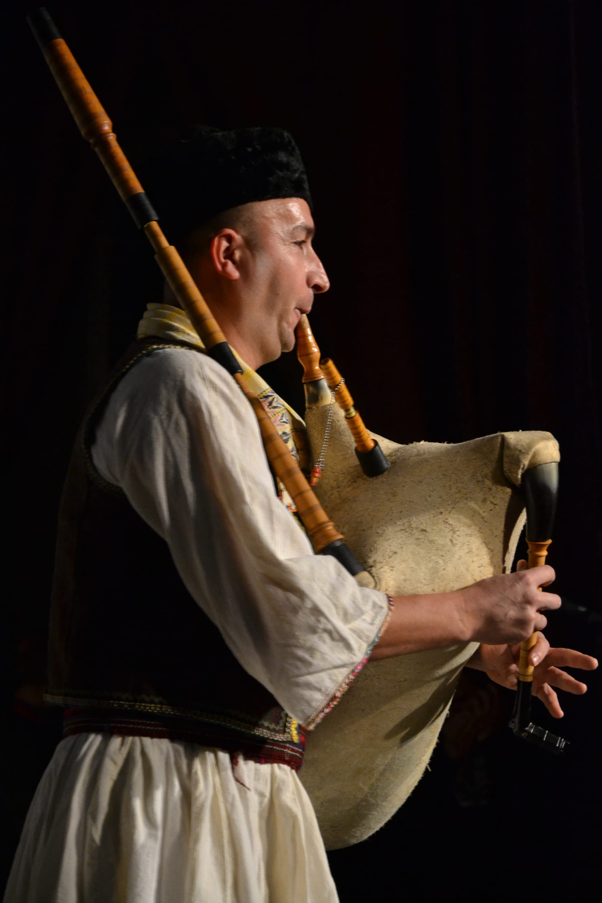 A man in a white shirt, dark embroidered waistcoat and fur hat plays a bagpipe with a pale goatskin bag and long wooden drone, against a dark background