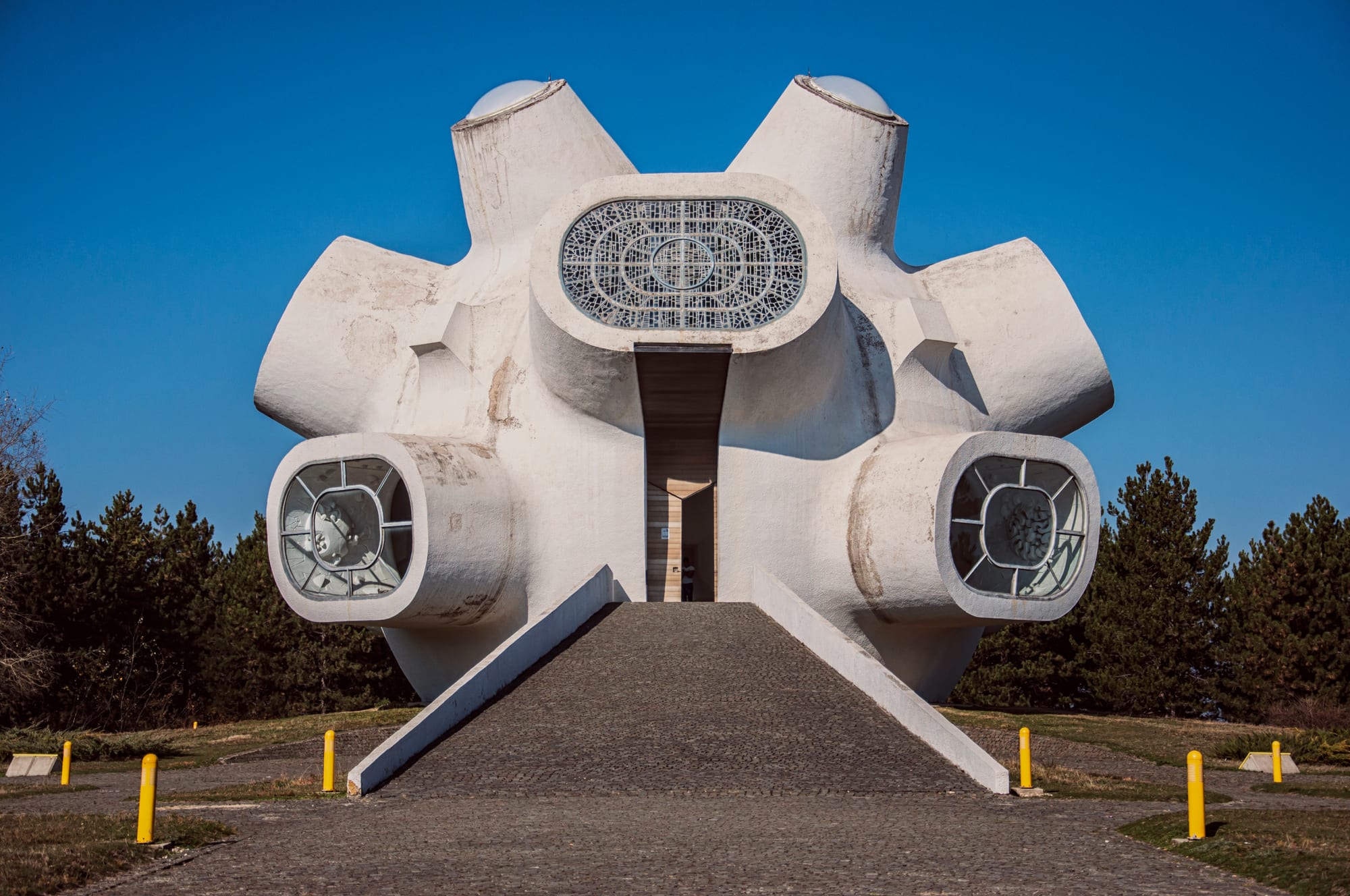 A white concrete monument shaped like a rounded body with protruding drum-like windows, reached by a paved ramp under a clear blue sky