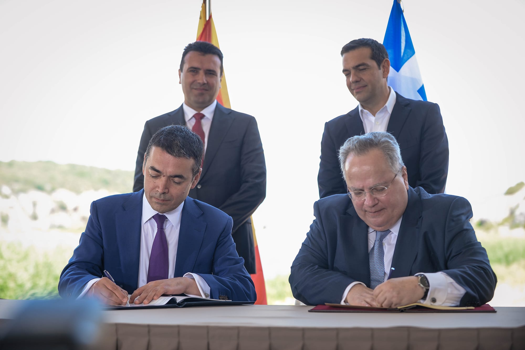 Two men in suits sign documents at a table while two others stand behind them in front of the flags of North Macedonia and Greece