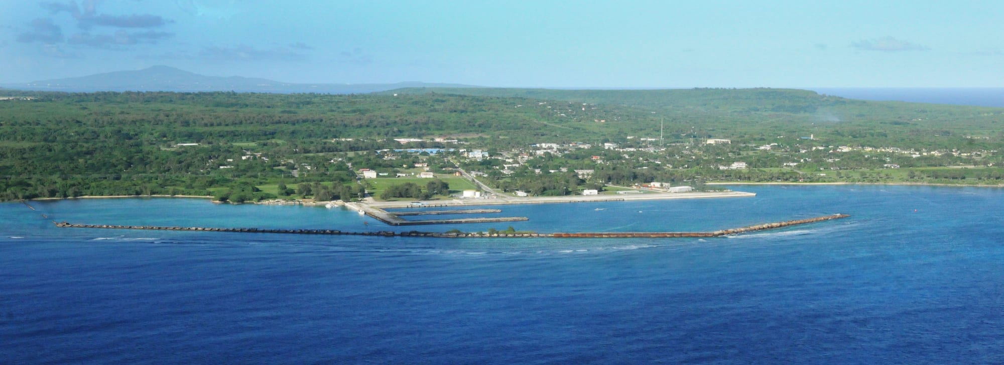 An aerial view of a small coastal village with low buildings scattered among trees beside a turquoise harbour