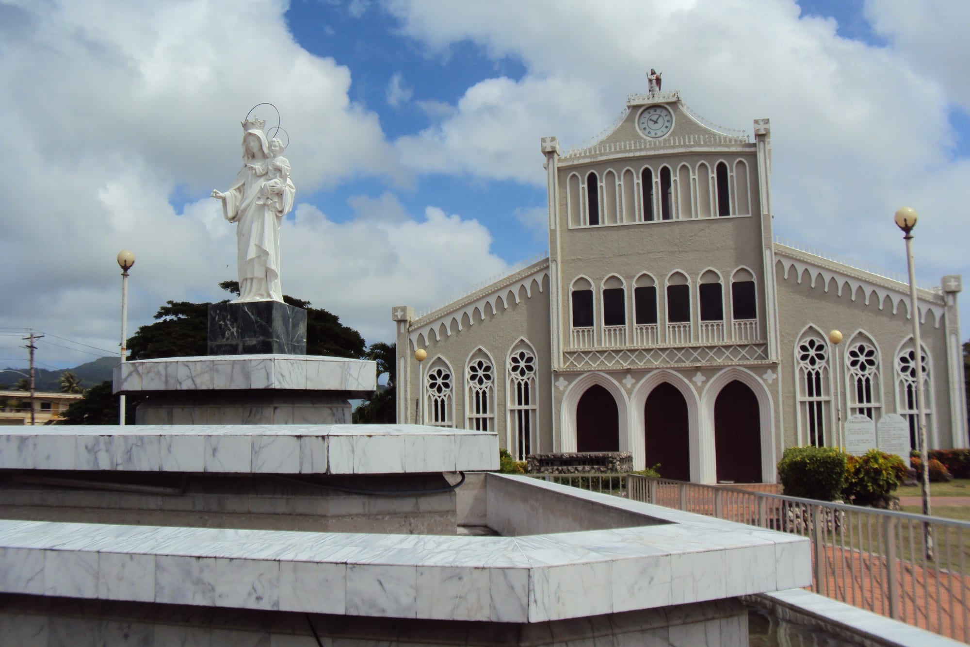 A white Gothic-revival Catholic cathedral with a statue of the Virgin Mary and child in front, under a blue sky