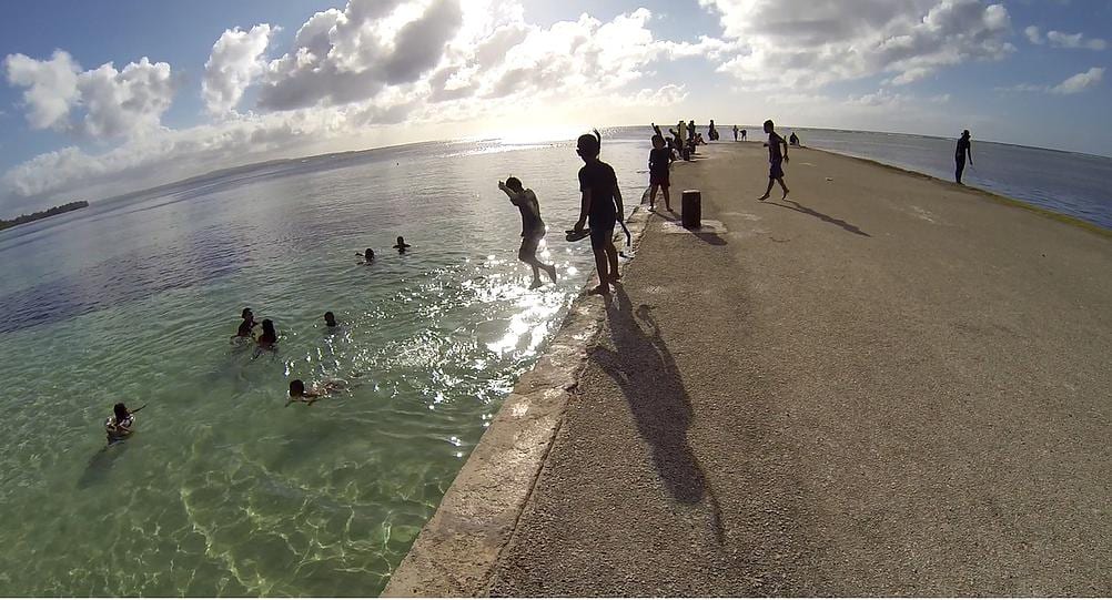 Children and teenagers swimming and jumping off a long concrete breakwater into clear turquoise water under a bright sky