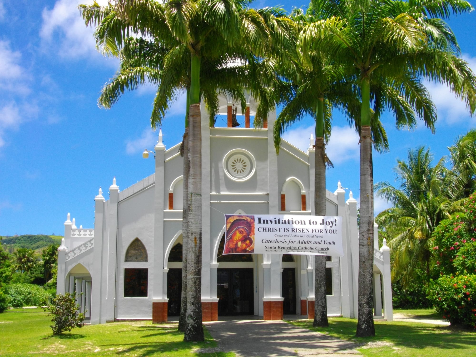A white Catholic church with a round window and bell tower, framed by tall royal palms, with a banner across the front showing an icon of the Virgin and Child