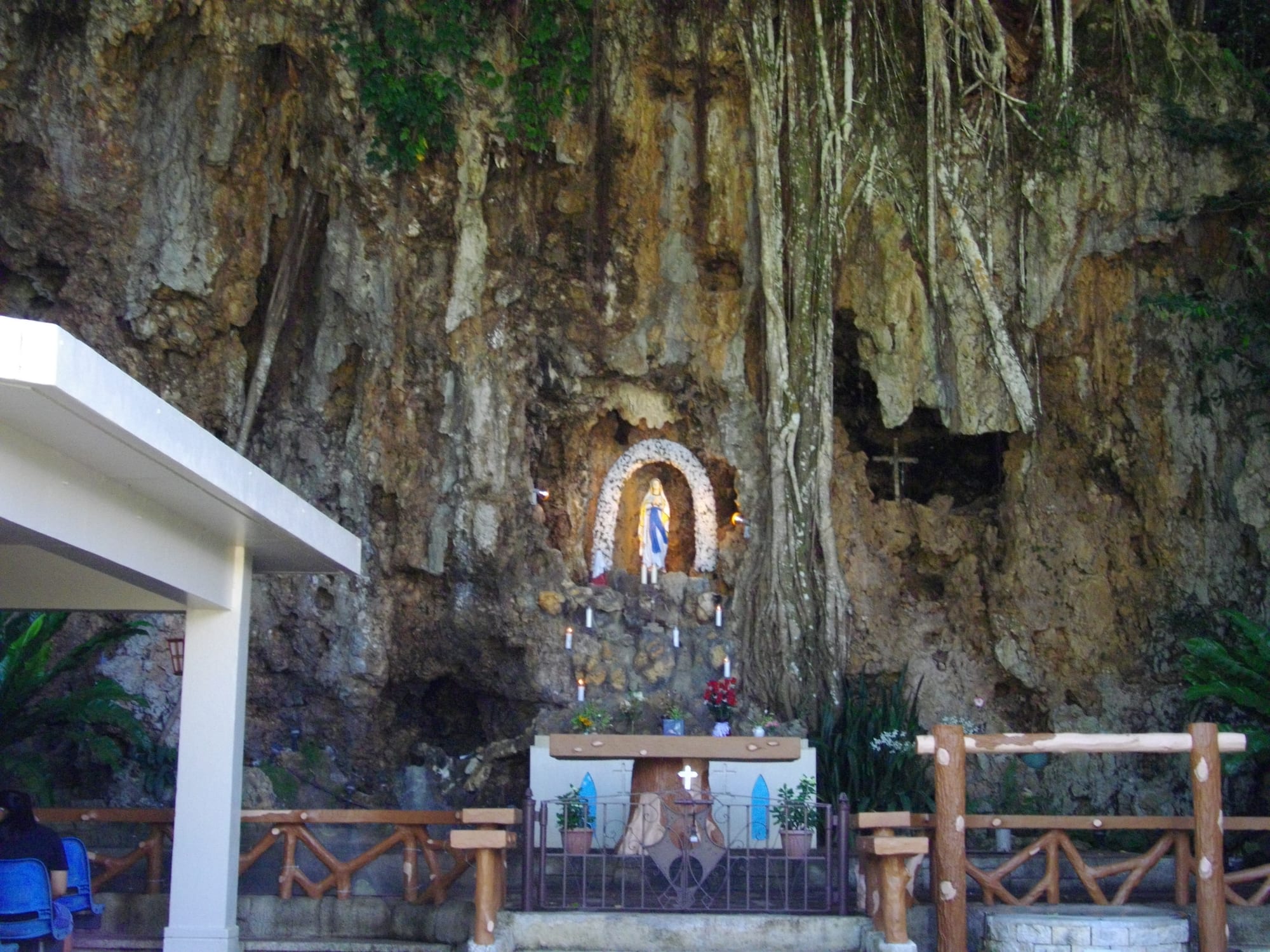 A white statue of the Virgin Mary in a flower-framed niche in a limestone cliff hung with tree roots, with lit candles on the rocks below, red flowers and a wooden altar table in front, and a covered seating area to the left