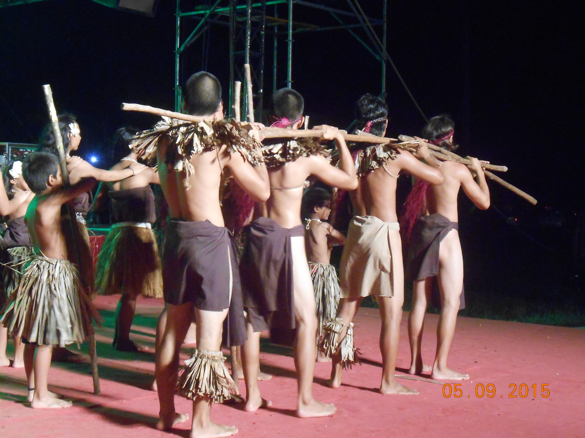 Performers seen from behind in fibre skirts and cloth wraps, some with woven shoulder pieces, carrying long wooden staves across their shoulders on a red stage at night