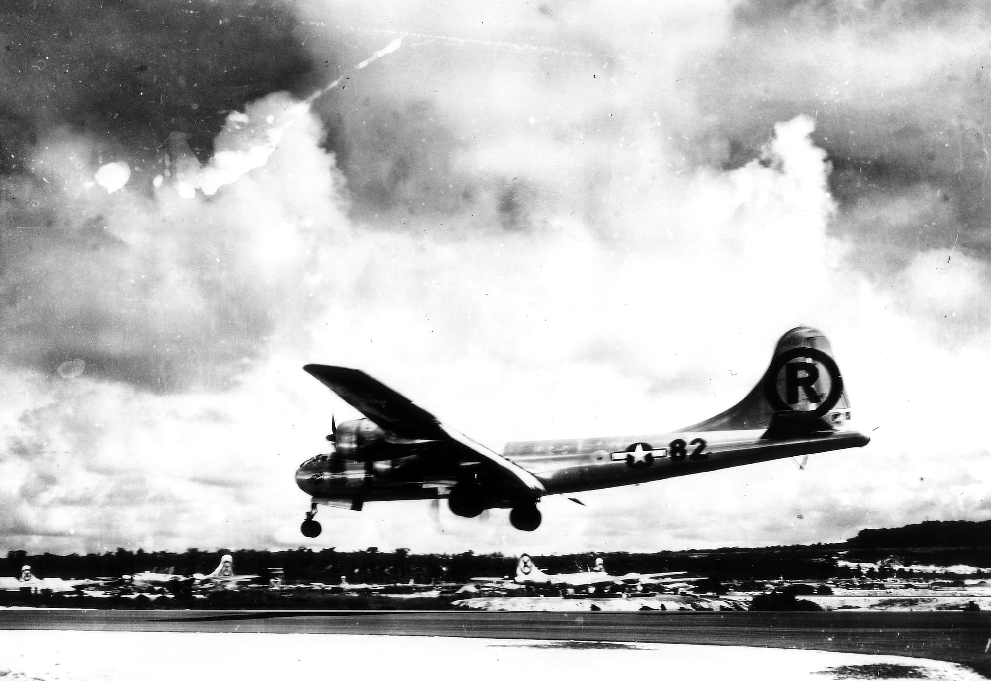 A black-and-white photograph of a four-engined B-29 bomber with a circled R on its tail, wheels down, just above a runway with other aircraft parked behind