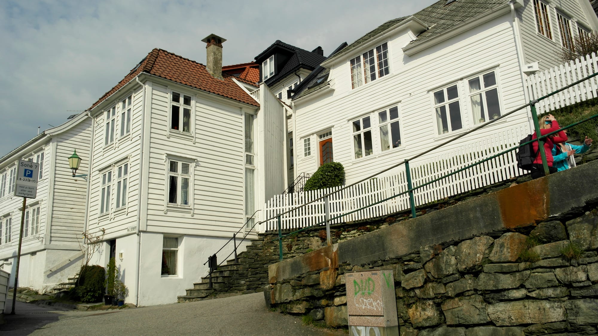 White wooden houses climbing a hillside street in Bergen, with two people walking past a stone retaining wall