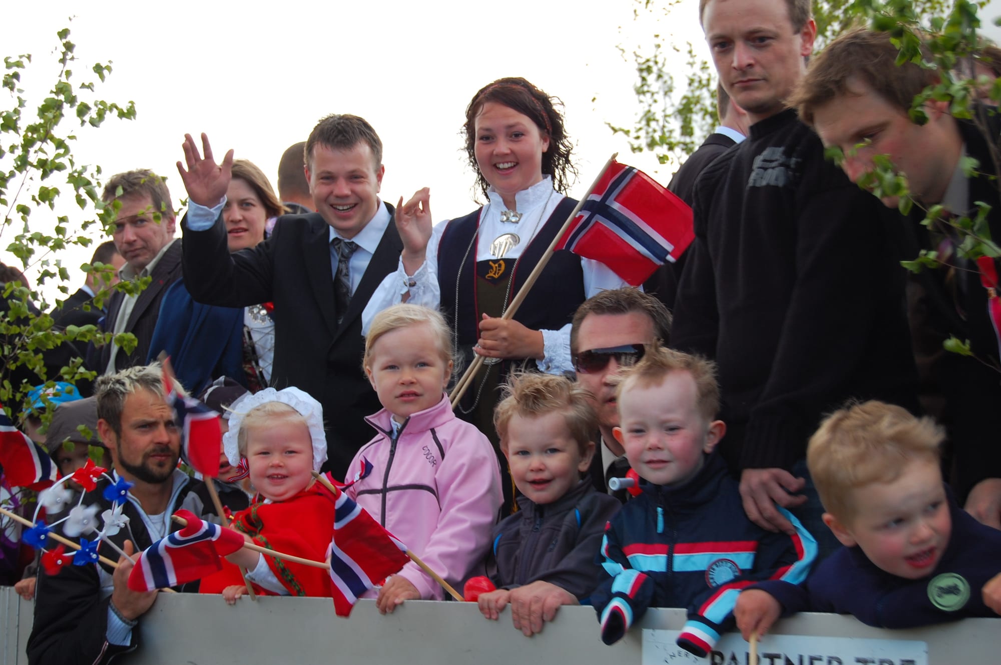 Kindergarten children and parents riding on the back of a decorated truck, some waving small Norwegian flags