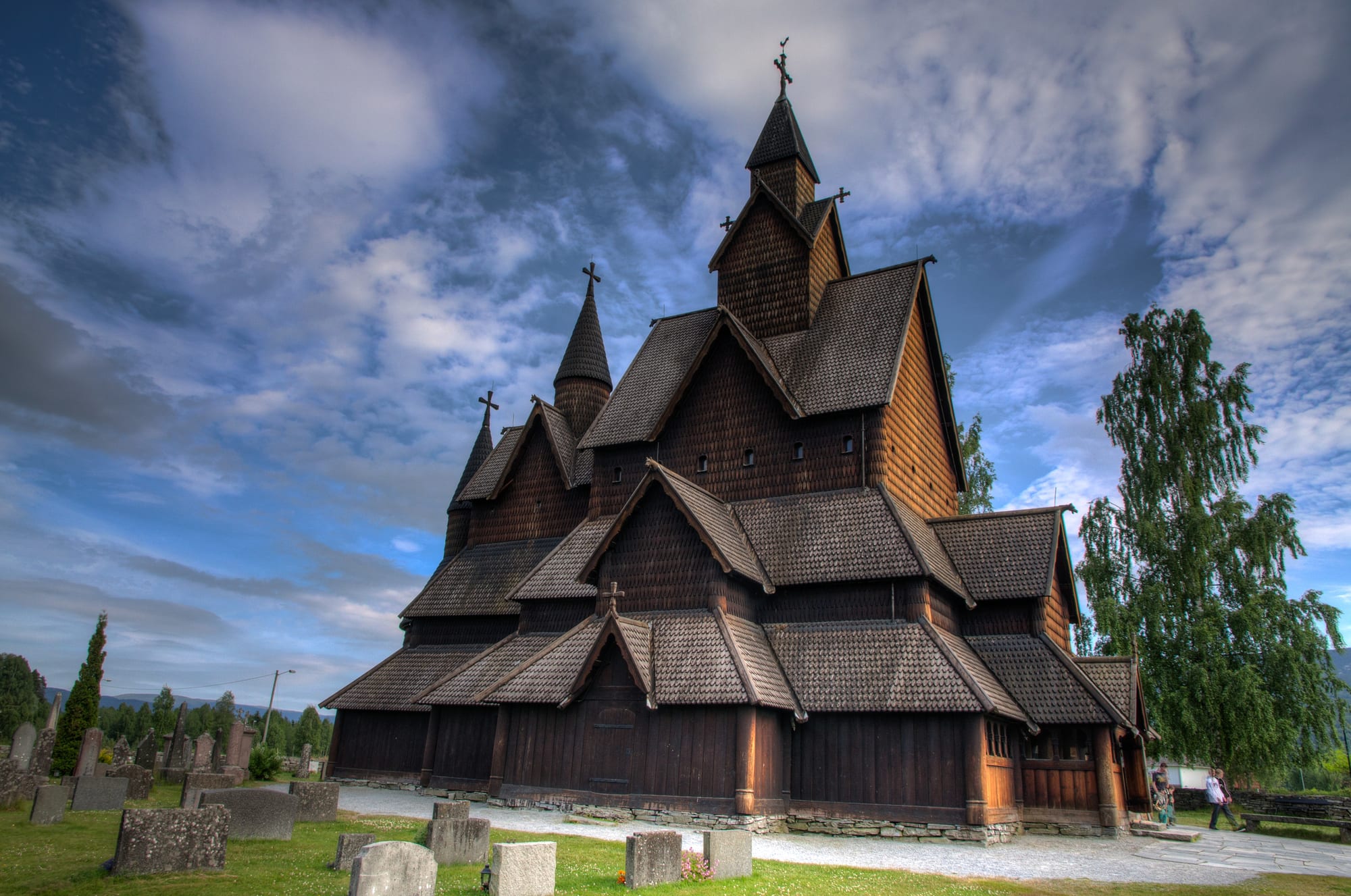 A large dark wooden stave church with steep tiered shingled roofs, small spires topped with crosses and a surrounding gallery, with gravestones in the grass in front