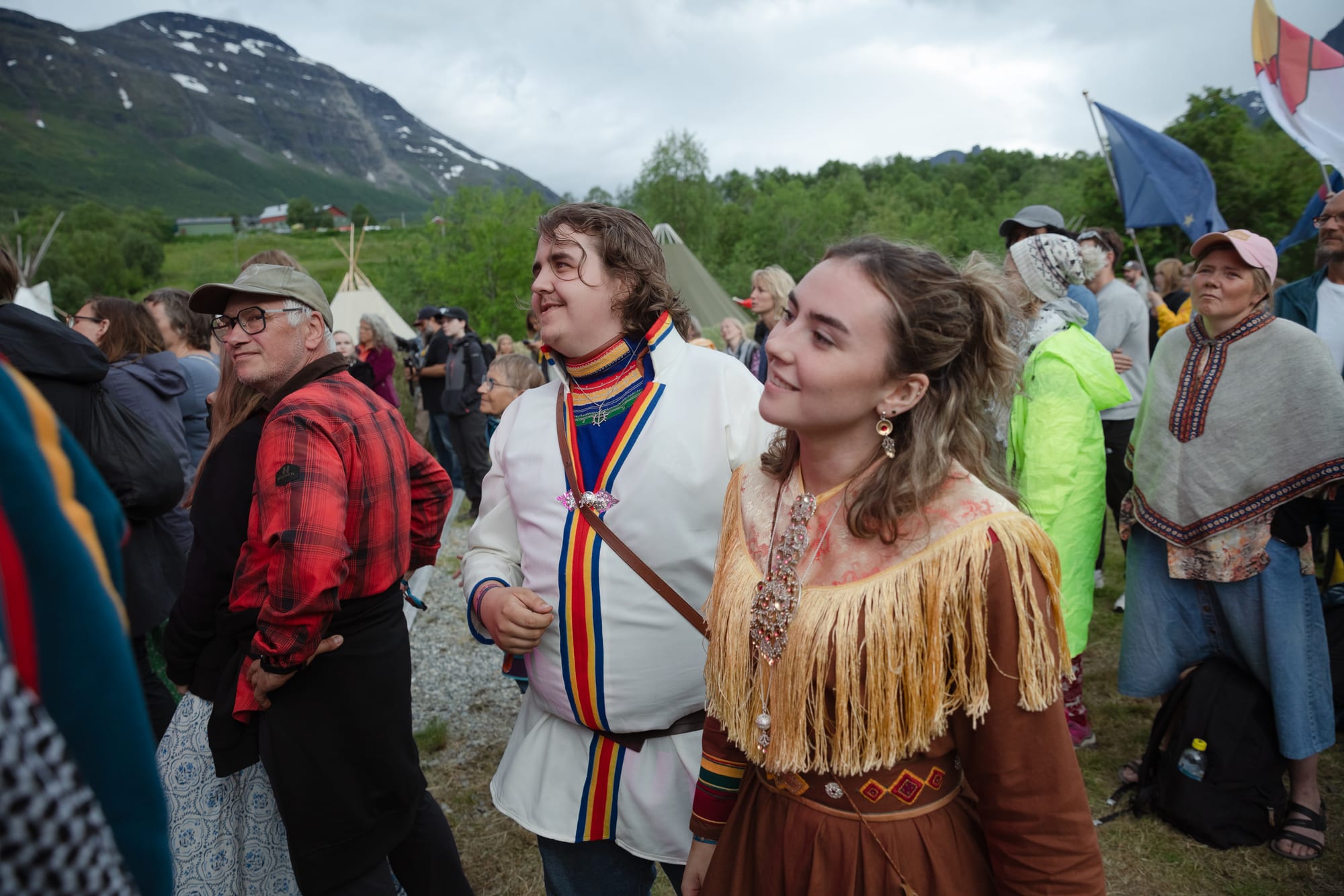 A crowd at an outdoor festival, with a young man in a white gákti trimmed in red, yellow and blue and a young woman in a brown gákti with a fringed shawl and silver jewellery, tents and mountains behind