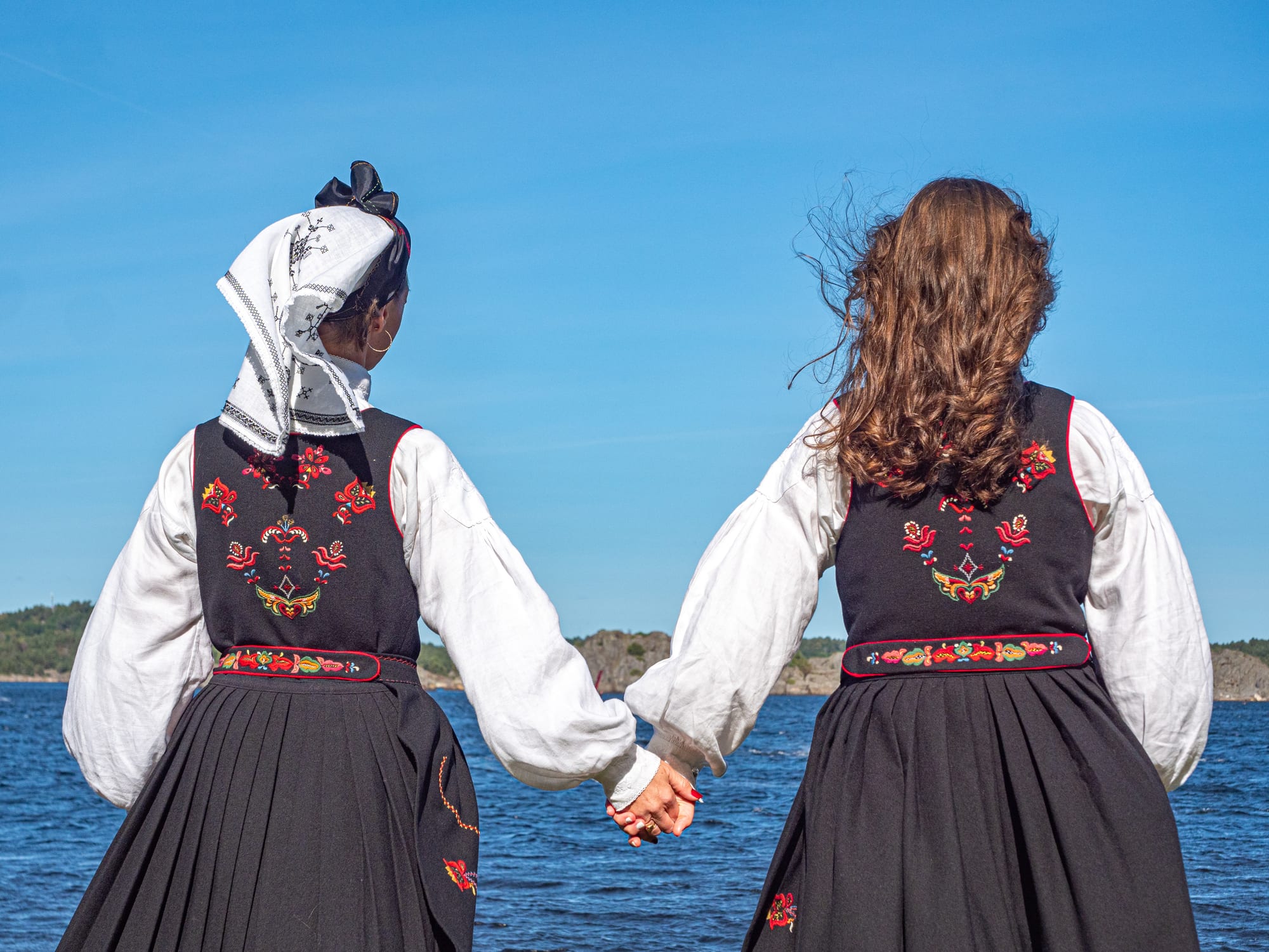 Two women seen from behind holding hands by the sea, both in black bunad skirts and bodices embroidered with red flowers, one wearing a white embroidered headscarf