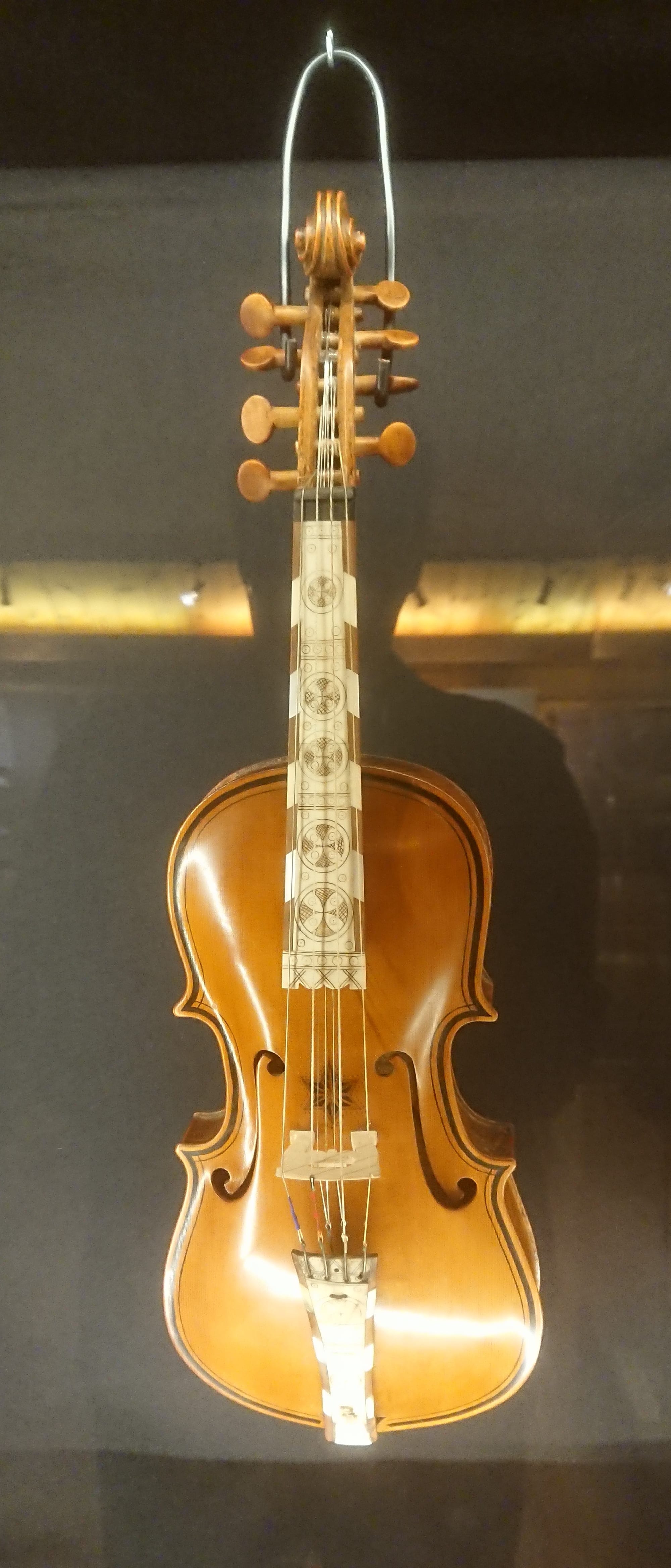 An ornate fiddle hanging in a display case, with a carved scroll, extra tuning pegs and a fingerboard inlaid with pale decorated panels