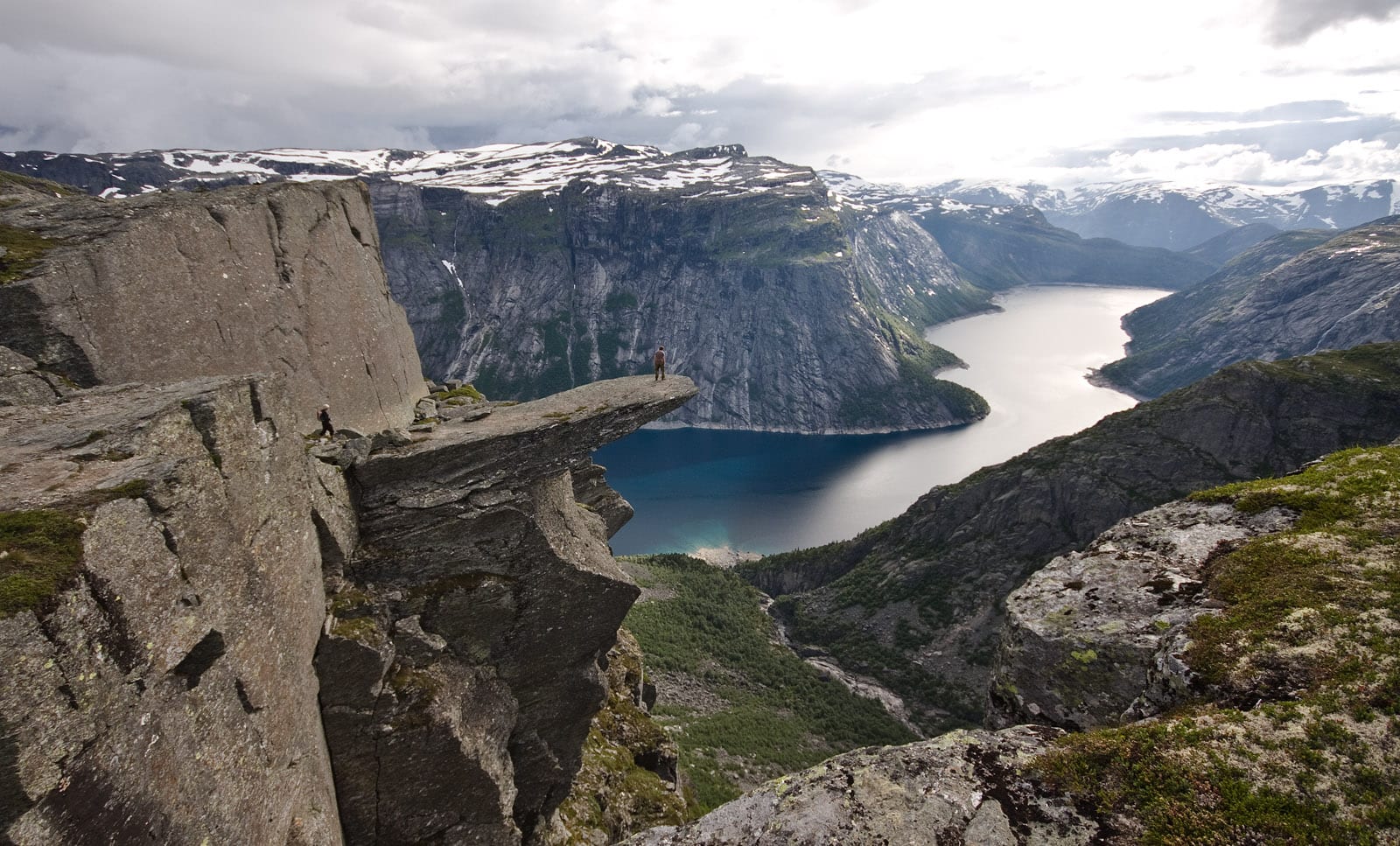 A person standing on a narrow horizontal rock ledge jutting out high above a lake