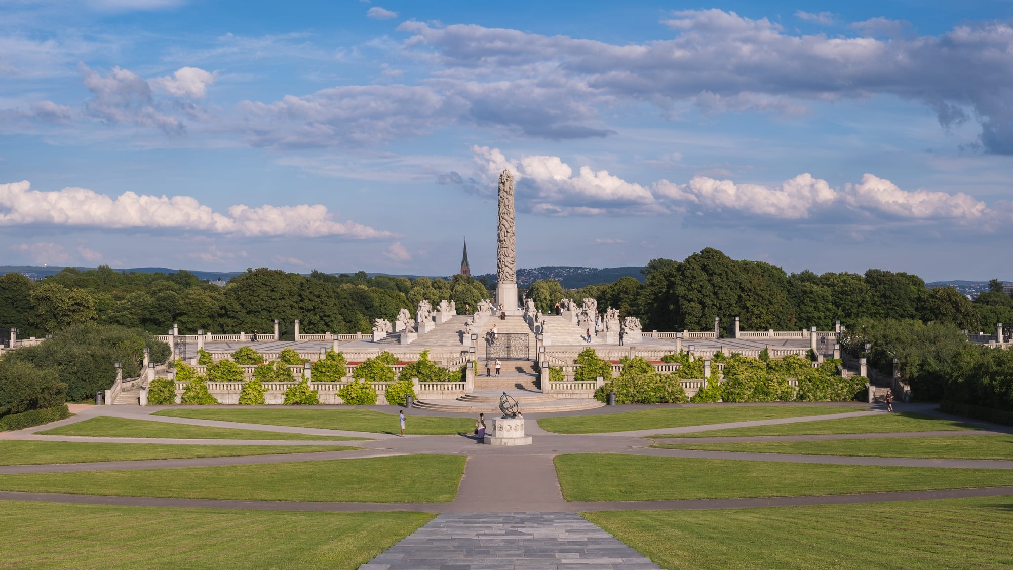 A wide park avenue lined with bronze statues leading to a tall stone monolith