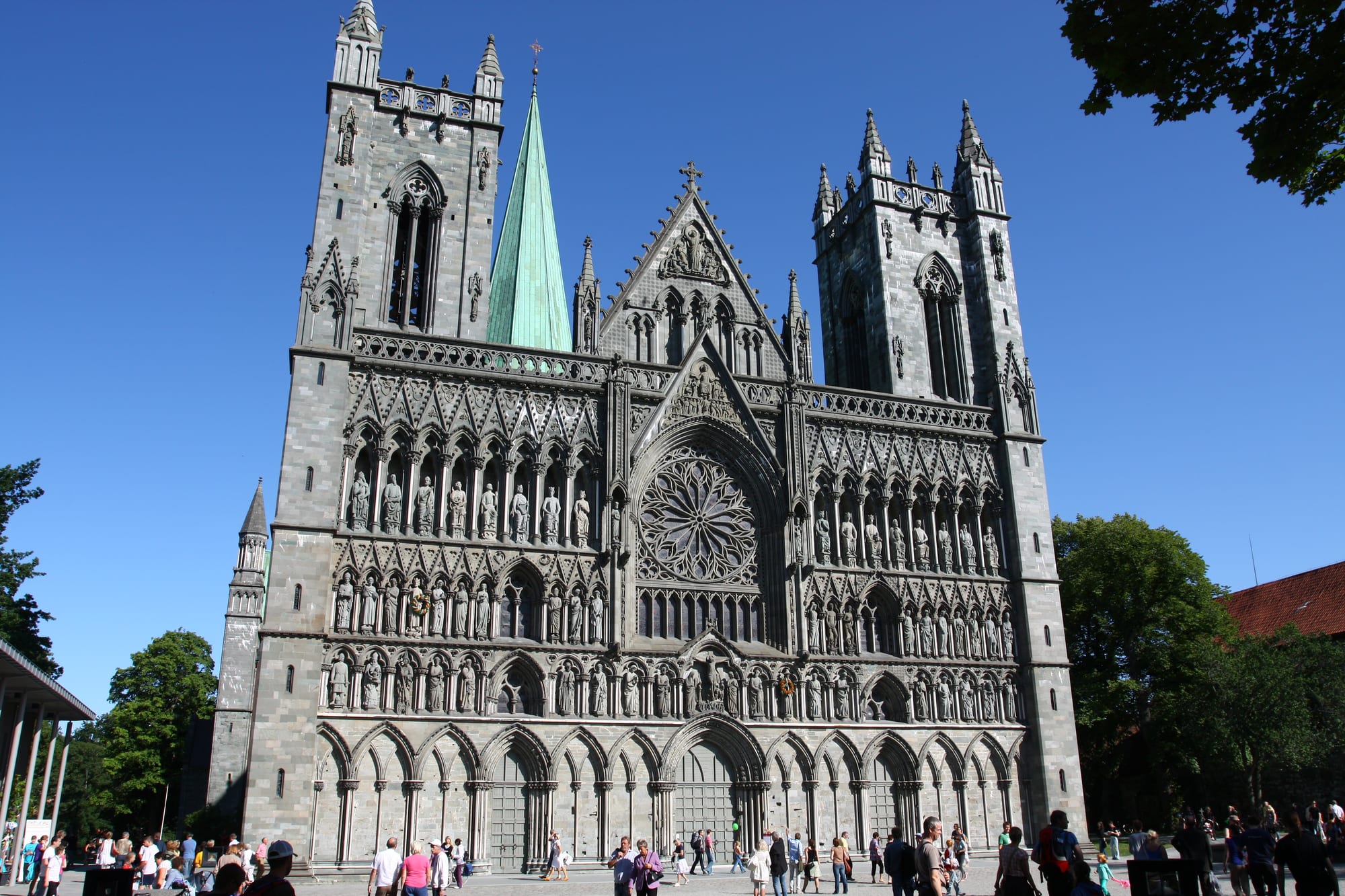 The grey stone Gothic west front of Nidaros Cathedral, with rows of carved statues, a rose window, two towers and a green copper spire under a clear blue sky
