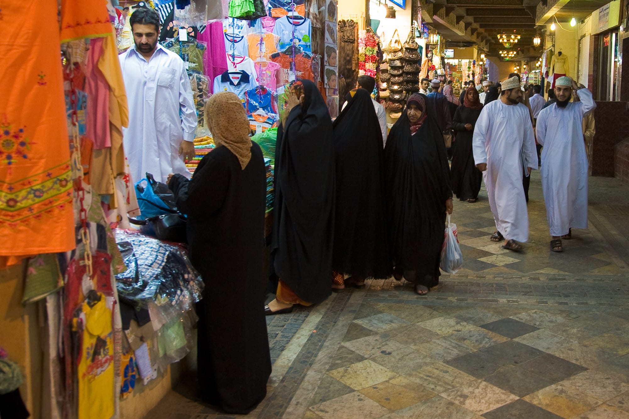 Shoppers in traditional Omani dress, women in black abaya and men in white dishdasha, walking through a covered souq lined with clothing stalls