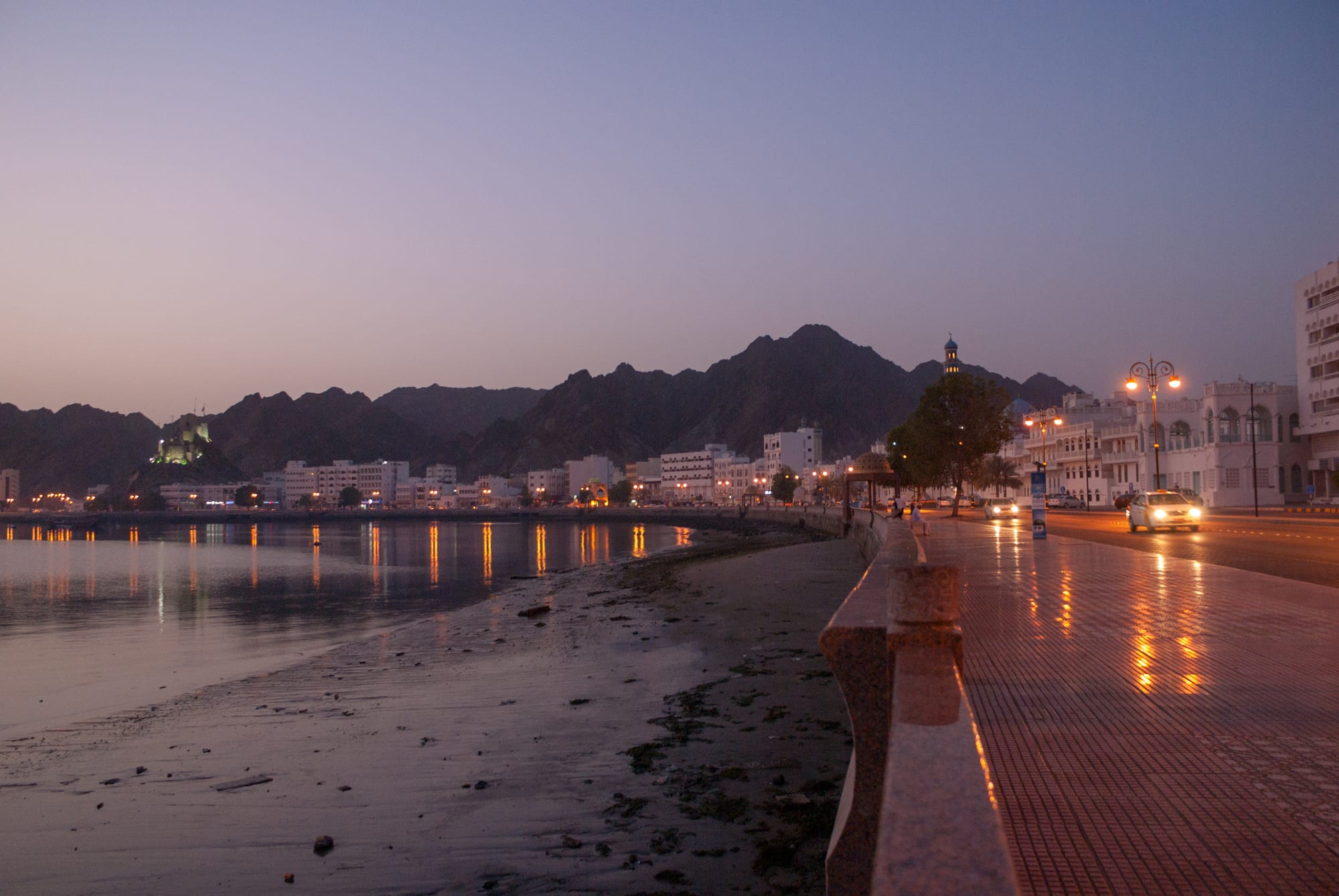 A waterfront promenade at dusk with lit buildings, a mosque minaret, mountains, and cars along the corniche road