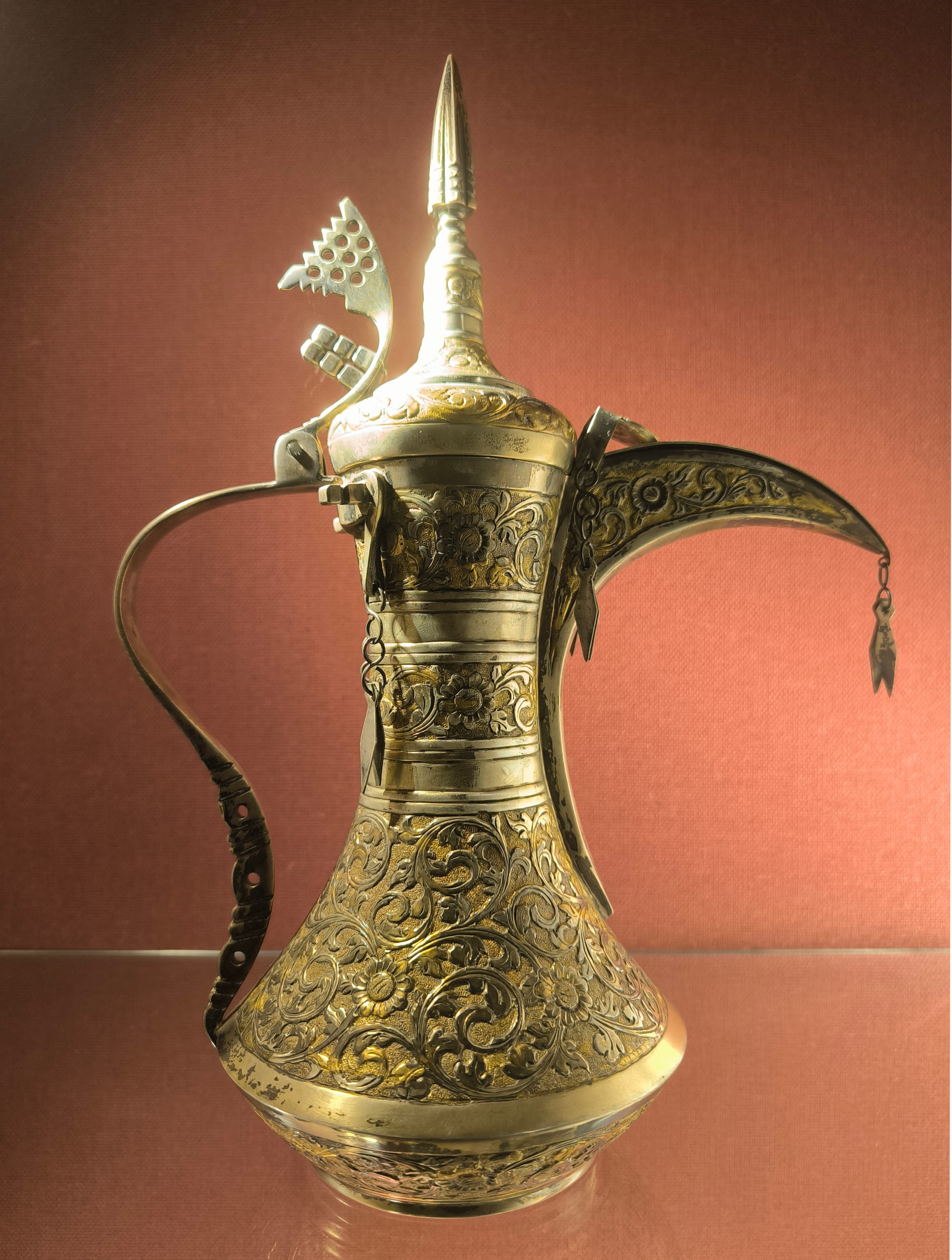 A tall engraved brass and copper coffee pot with a long curved spout, a pointed lid and an ornamental crest on the handle, displayed against a red background