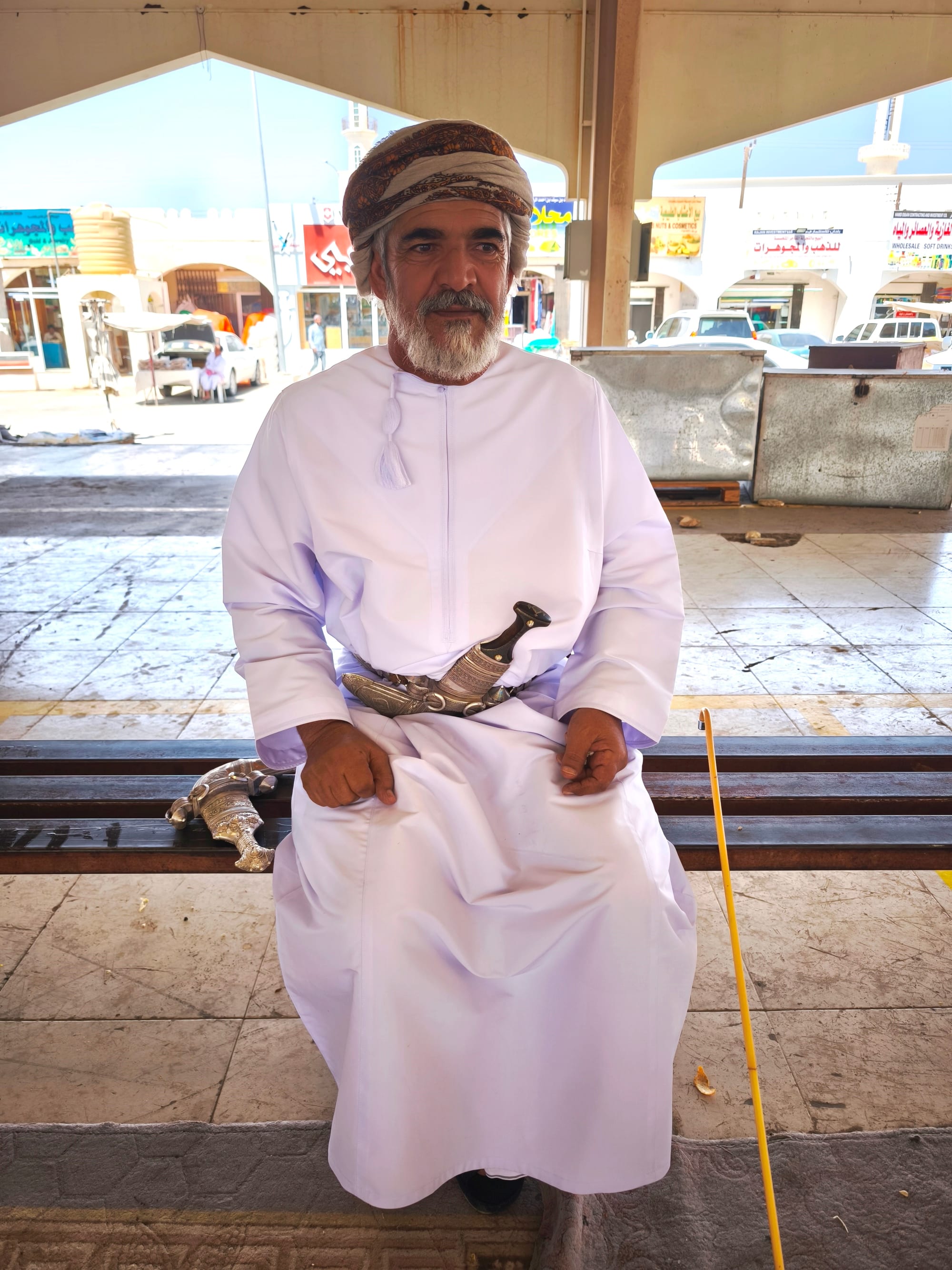 An older bearded man in a white dishdasha with a tassel at the collar and a patterned wrapped turban sits on a bench, a silver curved khanjar dagger at his waist