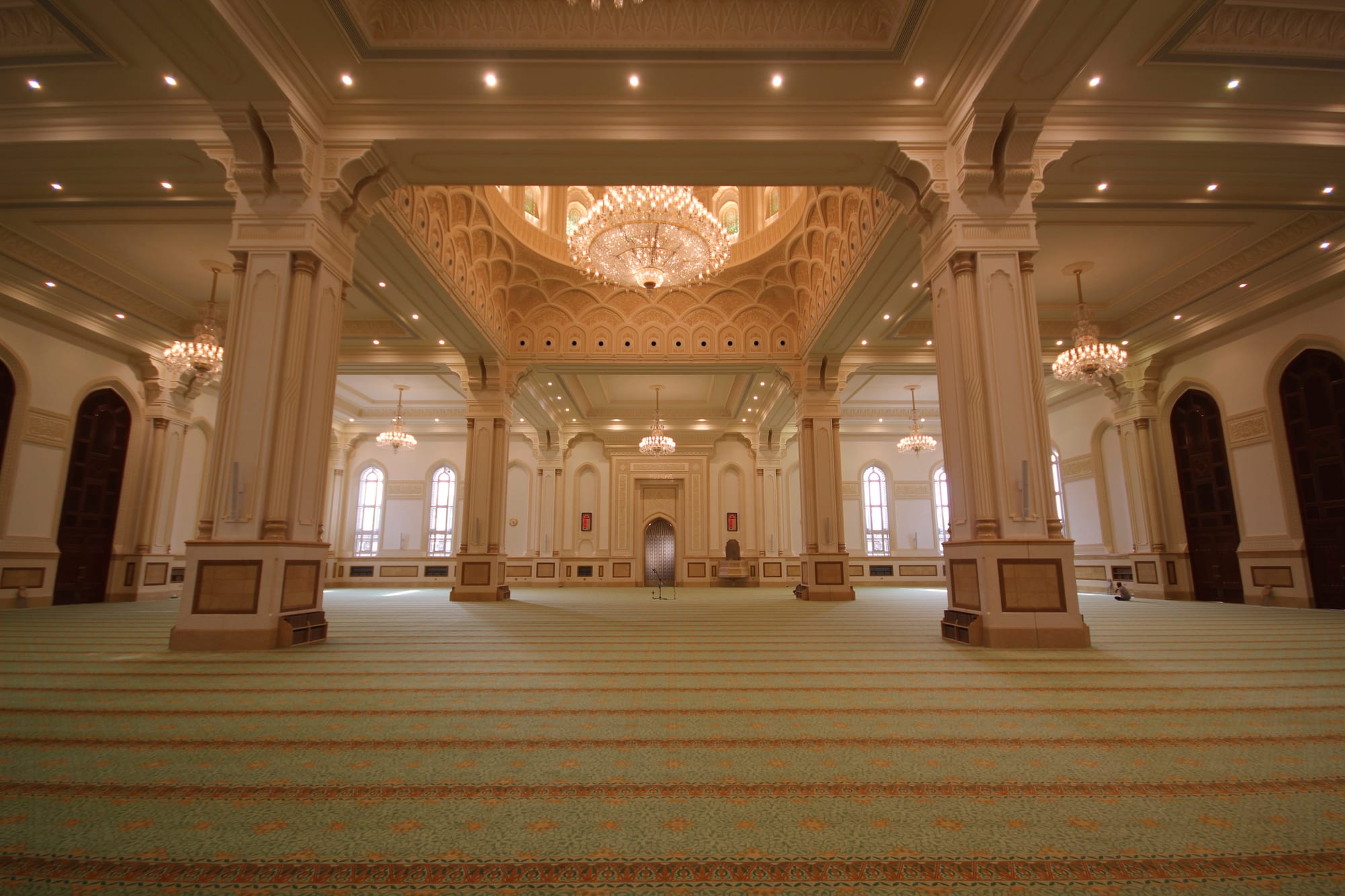 A vast, bright prayer hall with cream stone columns, chandeliers hanging from a decorated dome and a patterned green carpet stretching across the floor