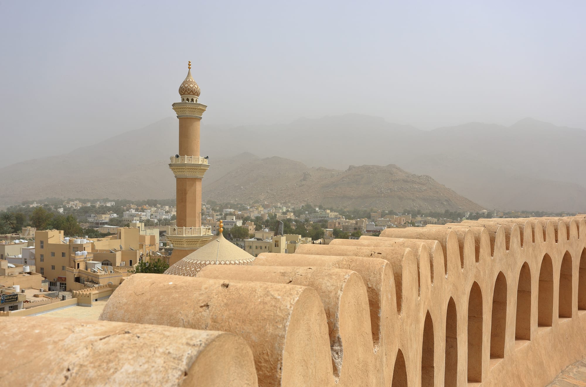 A massive round sandstone fortress tower beside a mosque minaret in the palm-ringed town of Nizwa