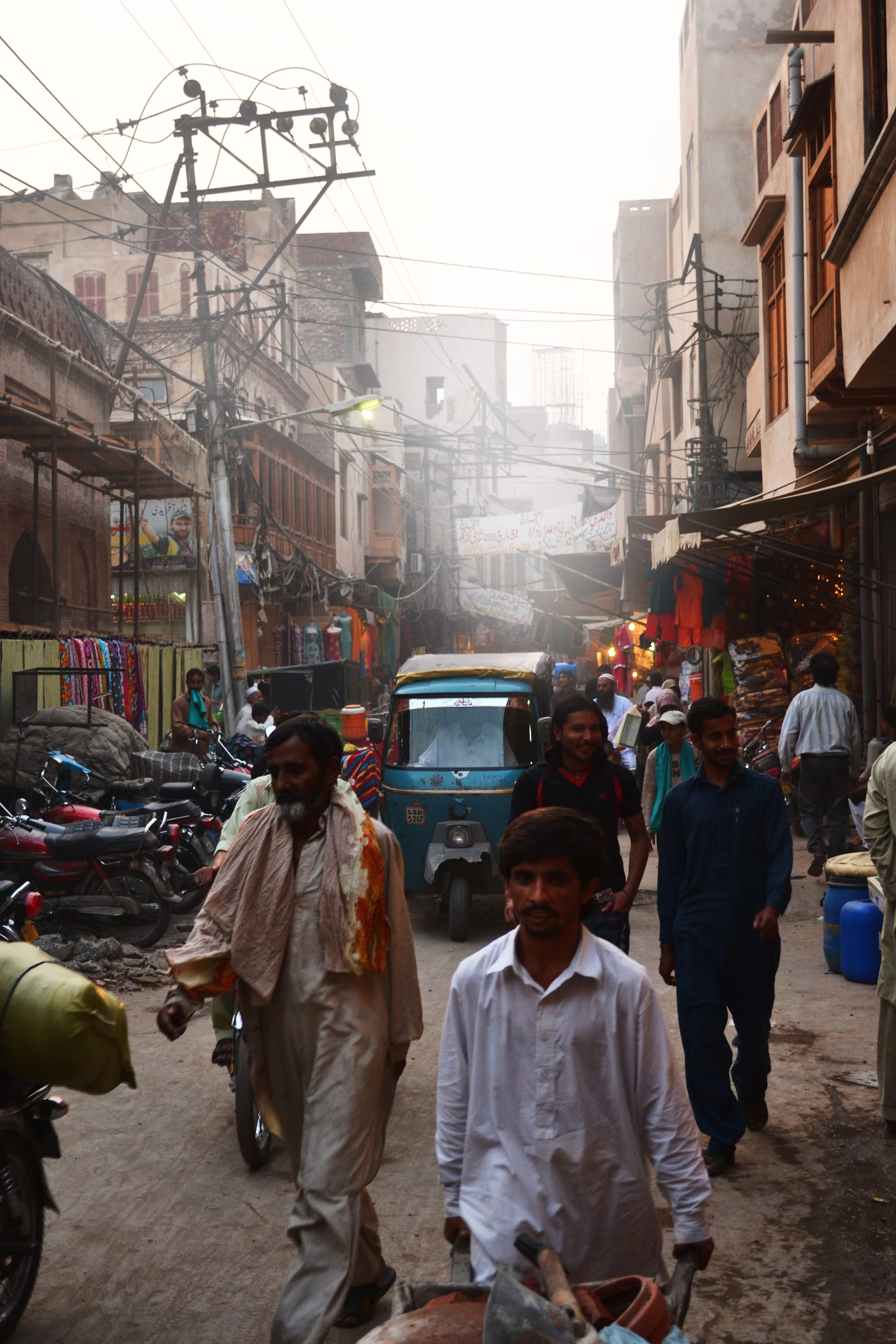 A crowded old-city street in Lahore with a rickshaw, motorbikes, pedestrians, overhead wires and shopfronts