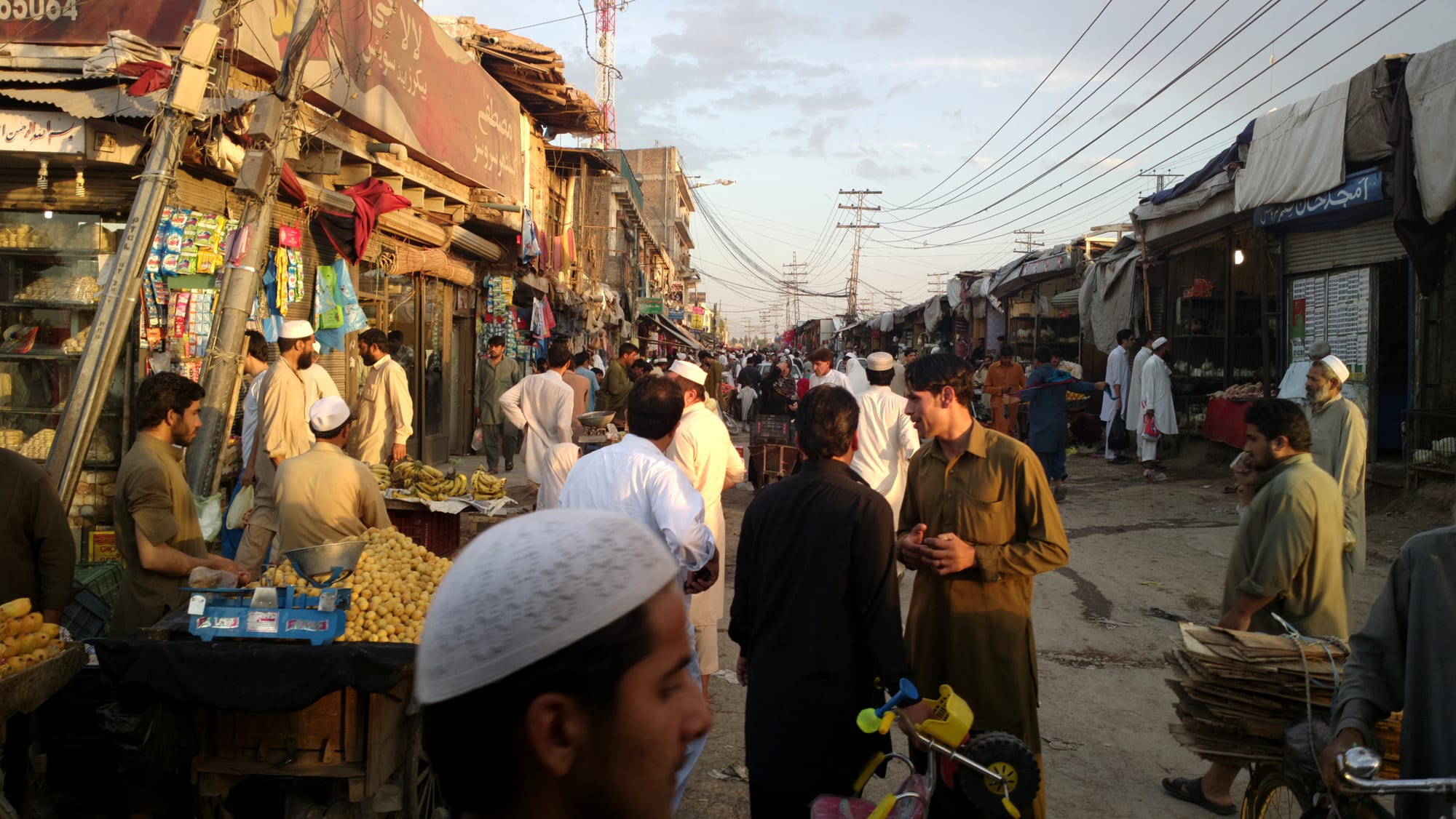 A crowded bazaar street in Peshawar at dusk, with fruit stalls, shoppers and vendors lining both sides