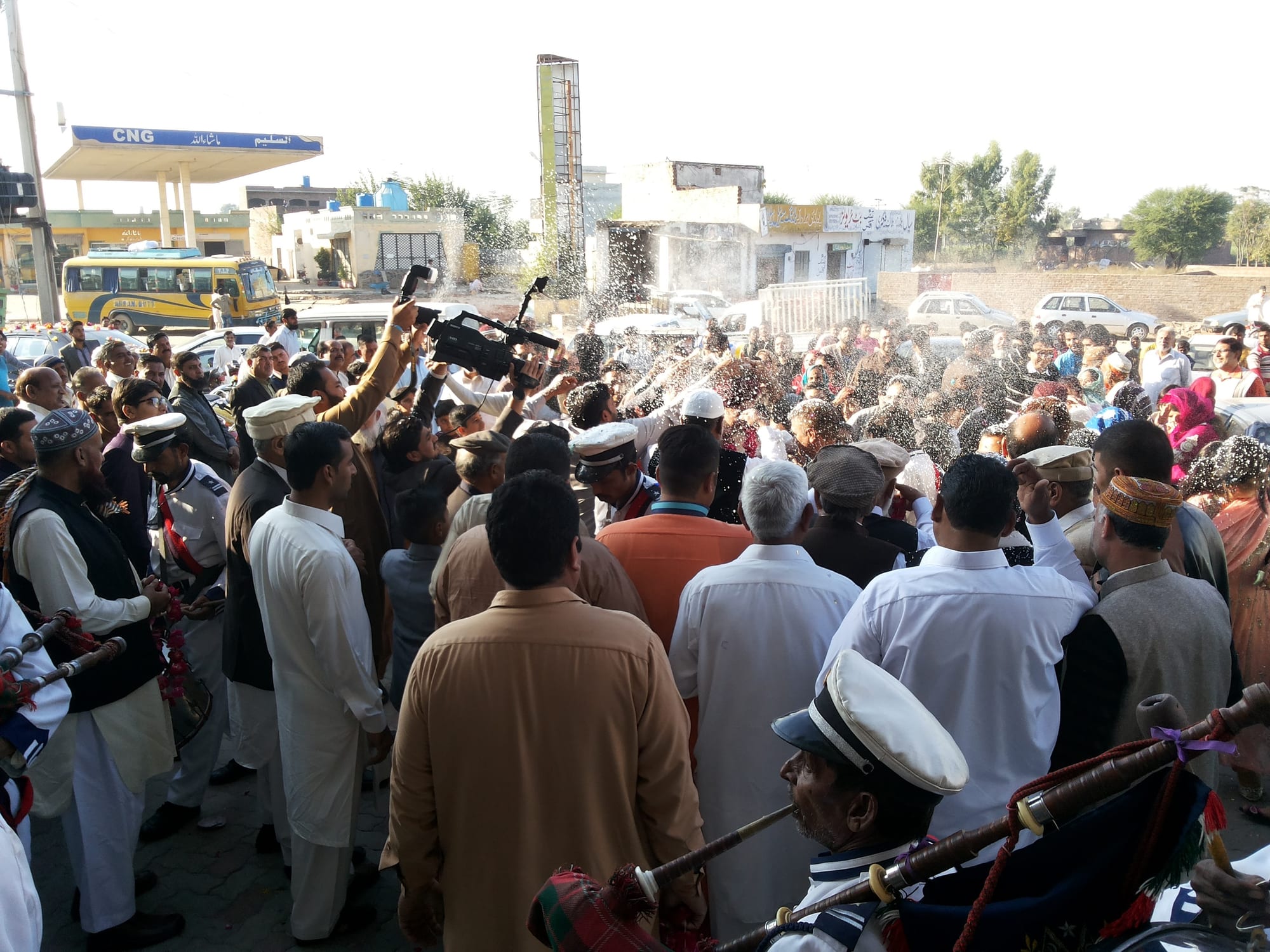 A dense crowd of men in shalwar kameez and caps gathers outside a building as flower petals fly through the air, while a man films on a video camera and pipers in white caps play in the foreground