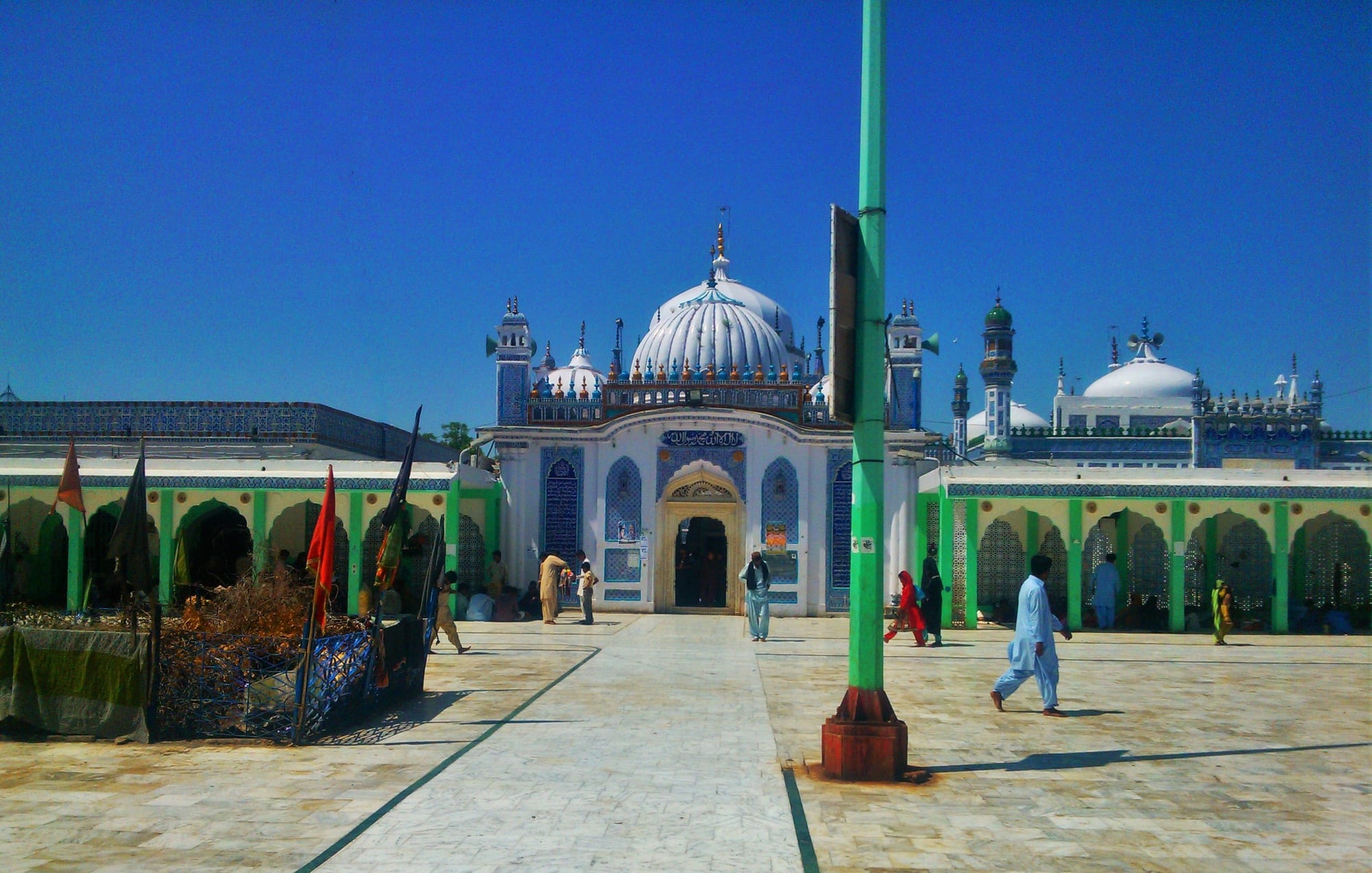A white-domed shrine with blue tiled walls and slender towers stands at the end of a wide paved courtyard under a deep blue sky, with a few visitors walking across it and flags on poles to one side