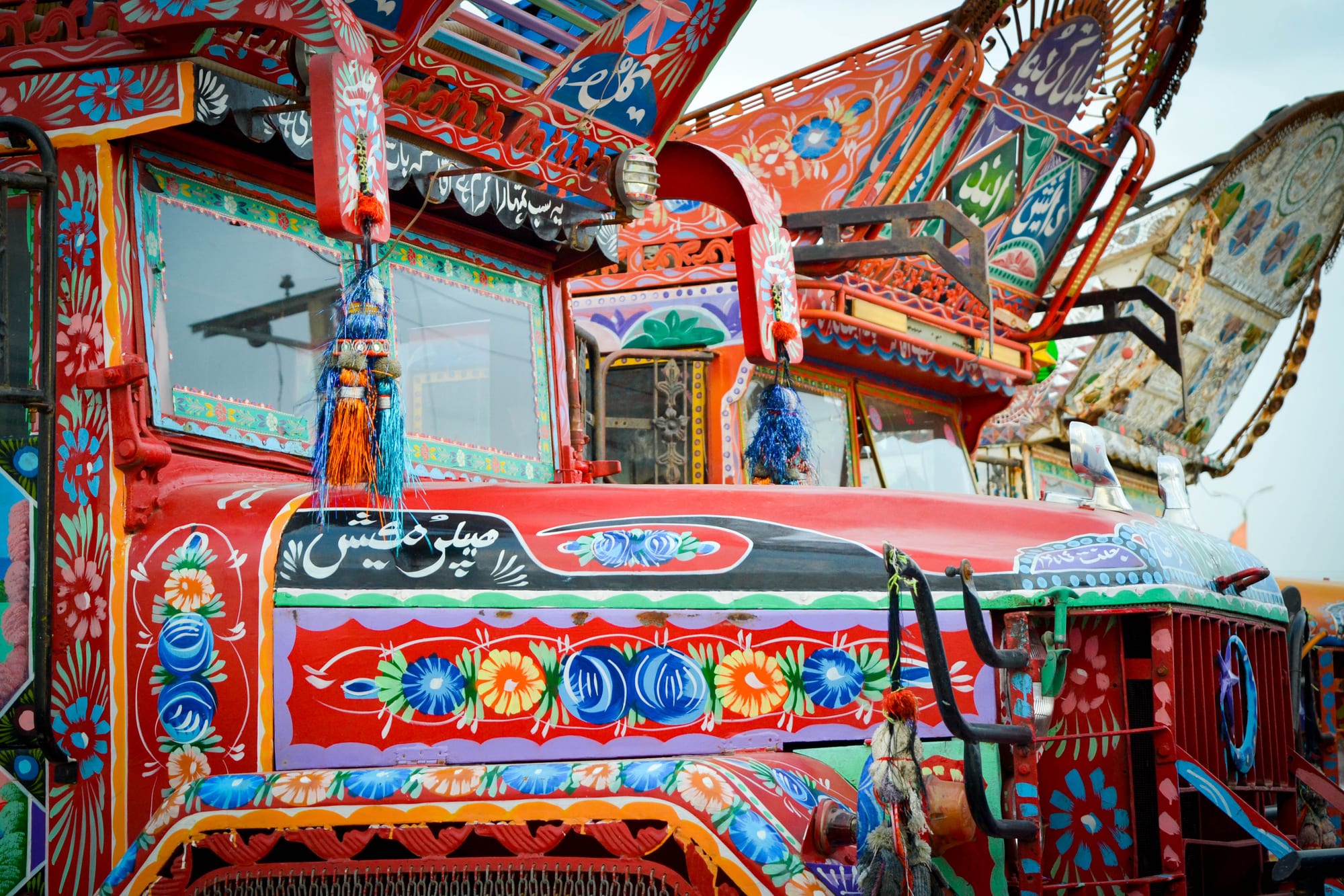 Close view of the front of brightly painted trucks covered in red, blue, orange and green floral patterns, with Urdu calligraphy panels, decorative metal fittings and blue and orange tassels hanging from the windscreen